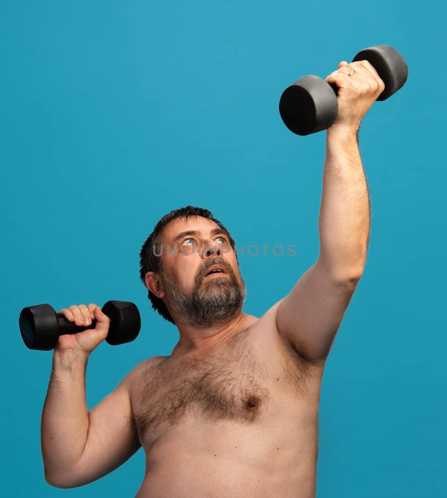 An elderly man without a shirt lifts dumbbells while exercising. His focused expression conveys determination. The background is a bright blue, enhancing the workout atmosphere.