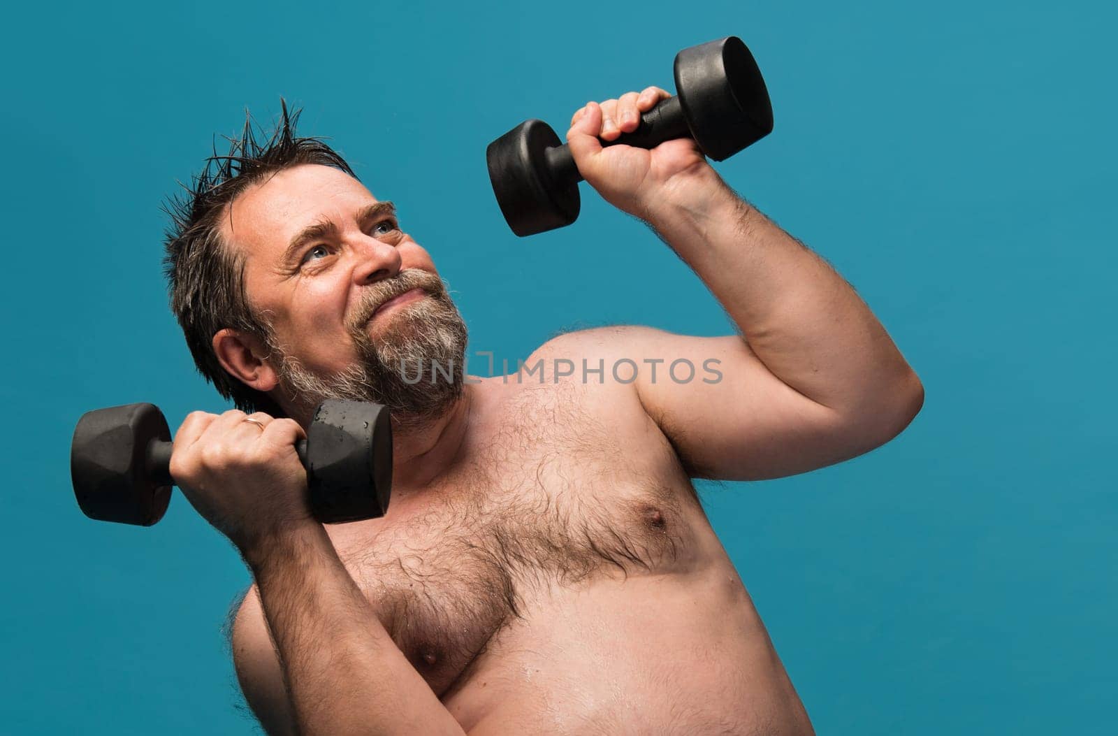 An elderly man is actively exercising with dumbbells in a bright blue fitness studio. He focuses on strength training while smiling, showcasing his dedication to fitness and well-being.