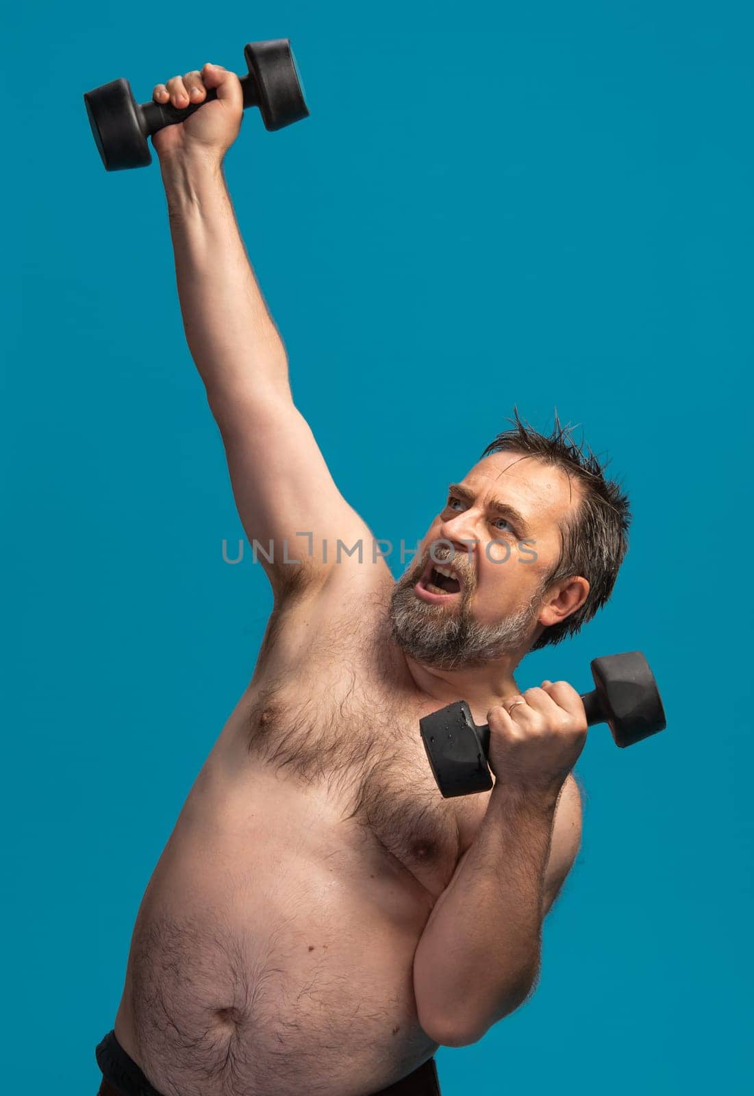An elderly man exercises vigorously with dumbbells, showcasing his commitment to fitness. He lifts one weight high while holding another weight at chest level, emphasizing strength training.