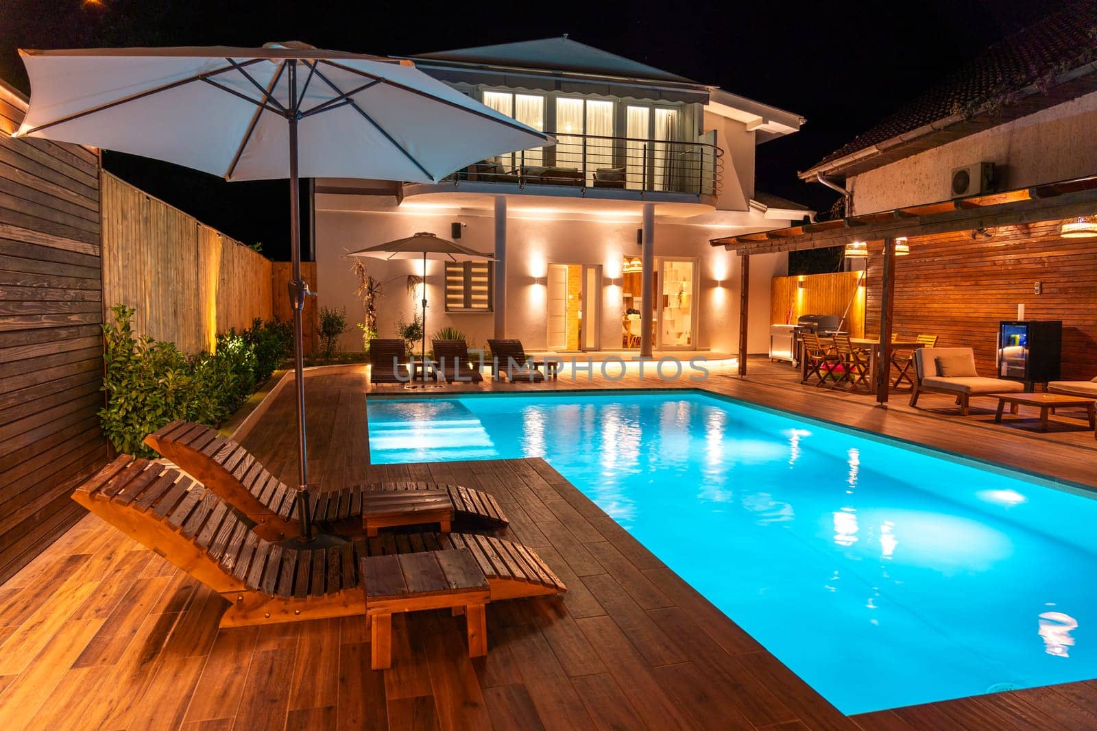 Modern house with a glowing swimming pool, comfortable lounge chairs, and warm lighting creating a relaxing backyard atmosphere. Concept of an ideal outdoor escape for leisure and comfort