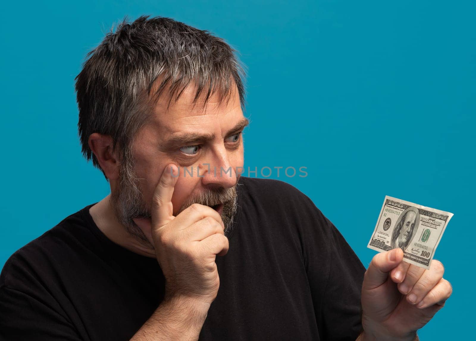 A man with gray hair and a beard thoughtfully examines a hundred-dollar bill while standing in front of a blue backdrop. His focus and expression convey curiosity and contemplation about the money.
