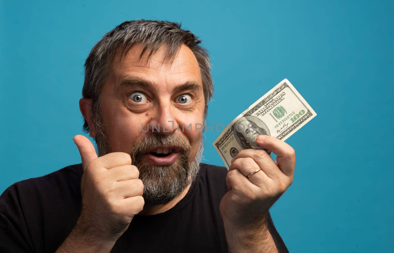 A man with a beard joyfully holds up a hundred-dollar bill while giving a thumbs up. His enthusiastic expression reflects happiness and excitement.