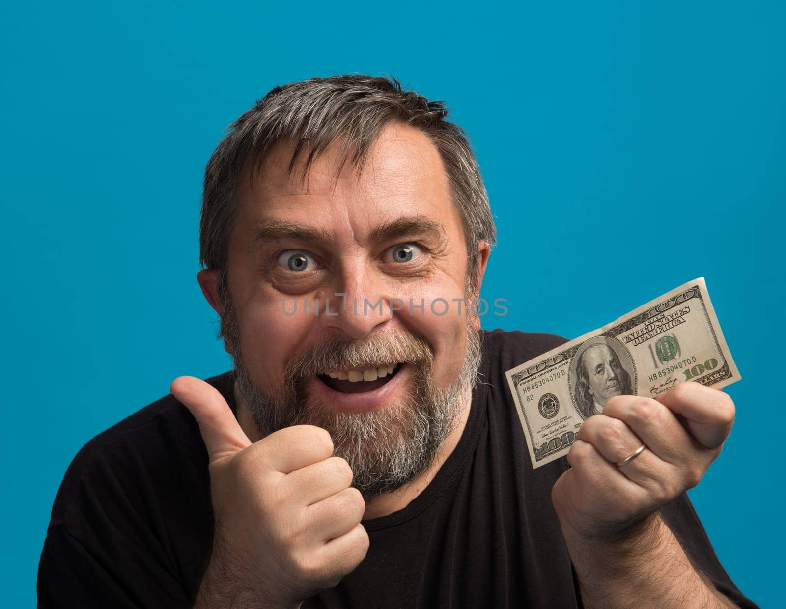 A man with a joyful expression holds a hundred-dollar bill in one hand while giving a thumbs-up with the other. The bright blue background enhances his enthusiastic demeanor.