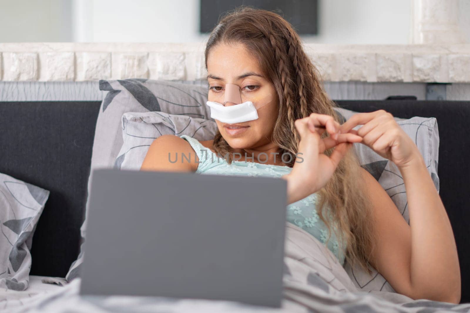 woman lies in bed in casual clothes and with bandages on her face after plastic surgery on nose, braids hair and watches TV on tablet,