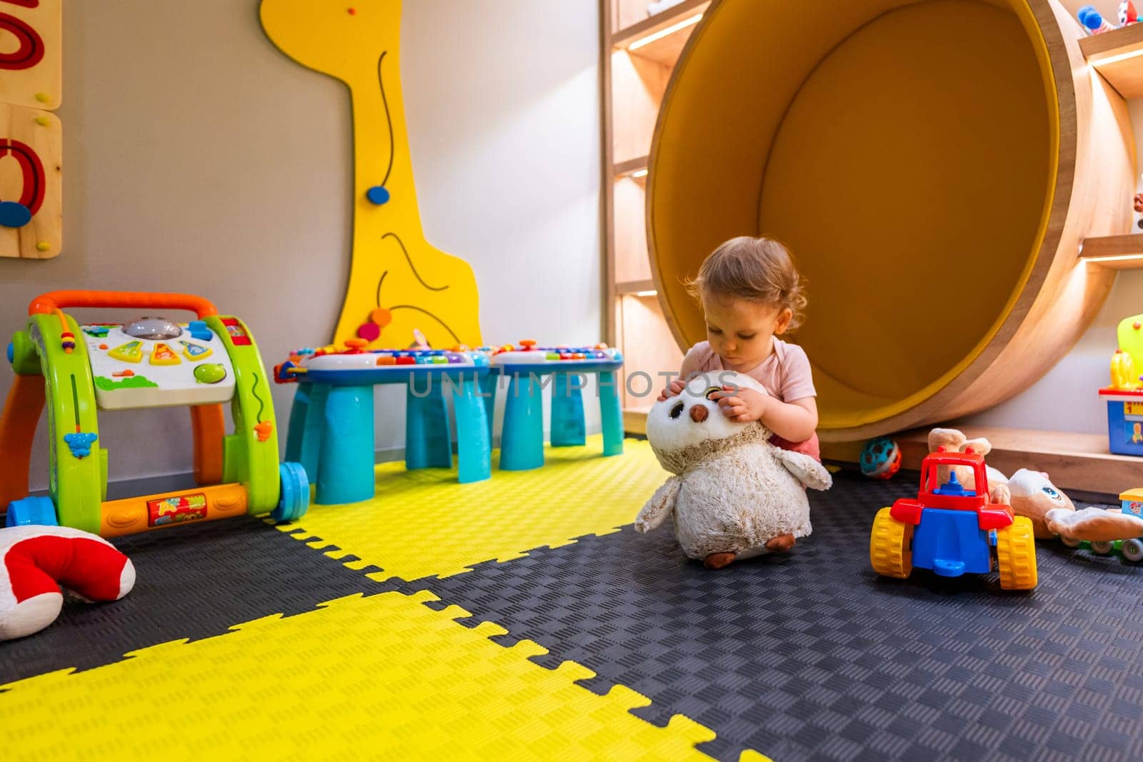 Young child sitting on soft play mats surrounded by toys and educational games in modern play area