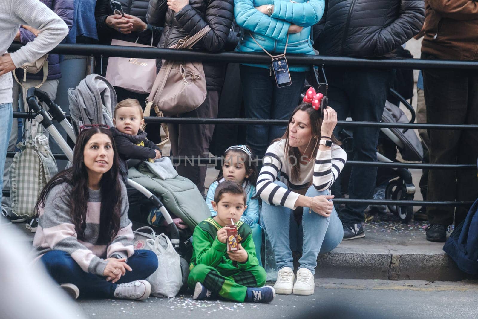 Family-friendly event spectators: Two mothers and three kids, one in a stroller, behind barriers at a public gathering. Crowded background indicates festive occasion.