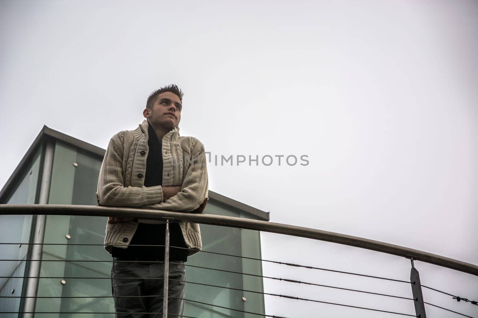 A man standing on a balcony with his arms crossed. Photo of a confident man standing on a balcony with crossed arms in a cloudy day in winter, seen from below