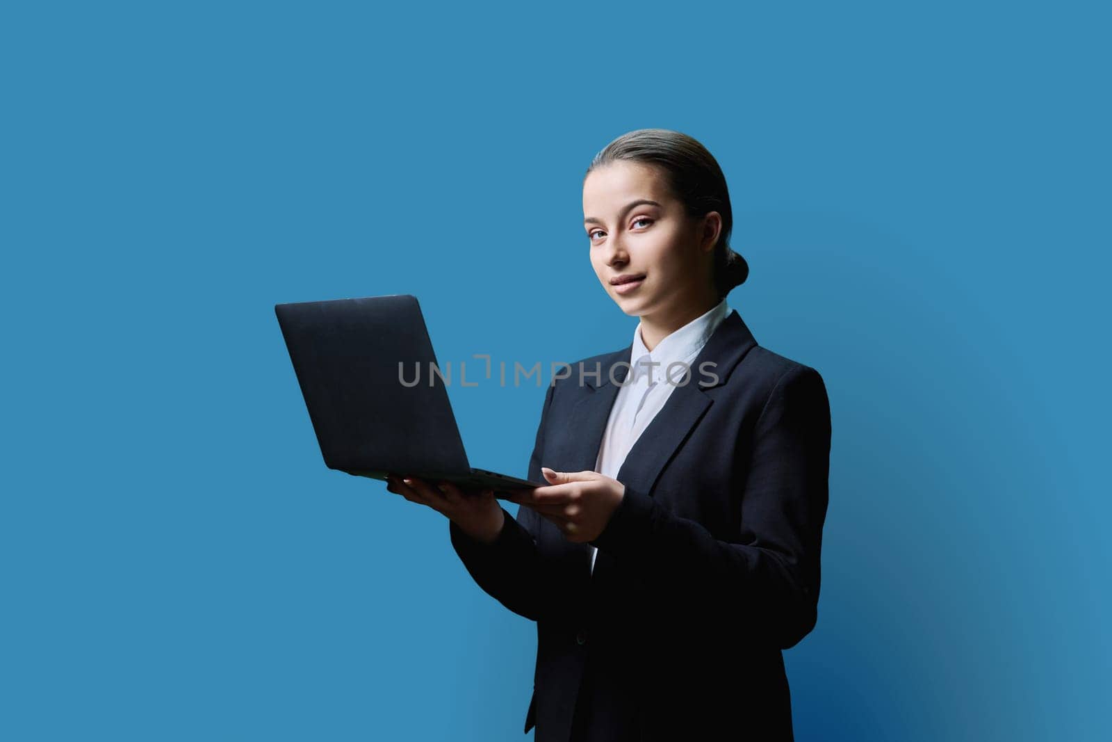 Teenage female student in formal business style using computer laptop, on blue studio background. Digital technologies, education, virtual educational services, online learning, youth concept