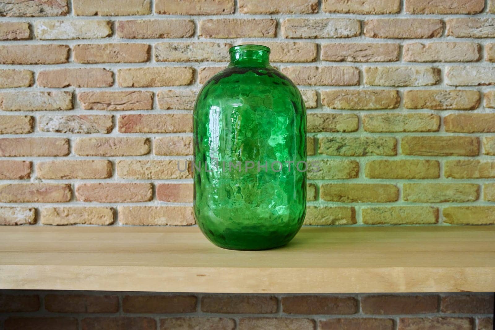 Green glass interior vase bottle on wooden desk, brick wall background. Background, copy space, design, style, interior, decor, house concept