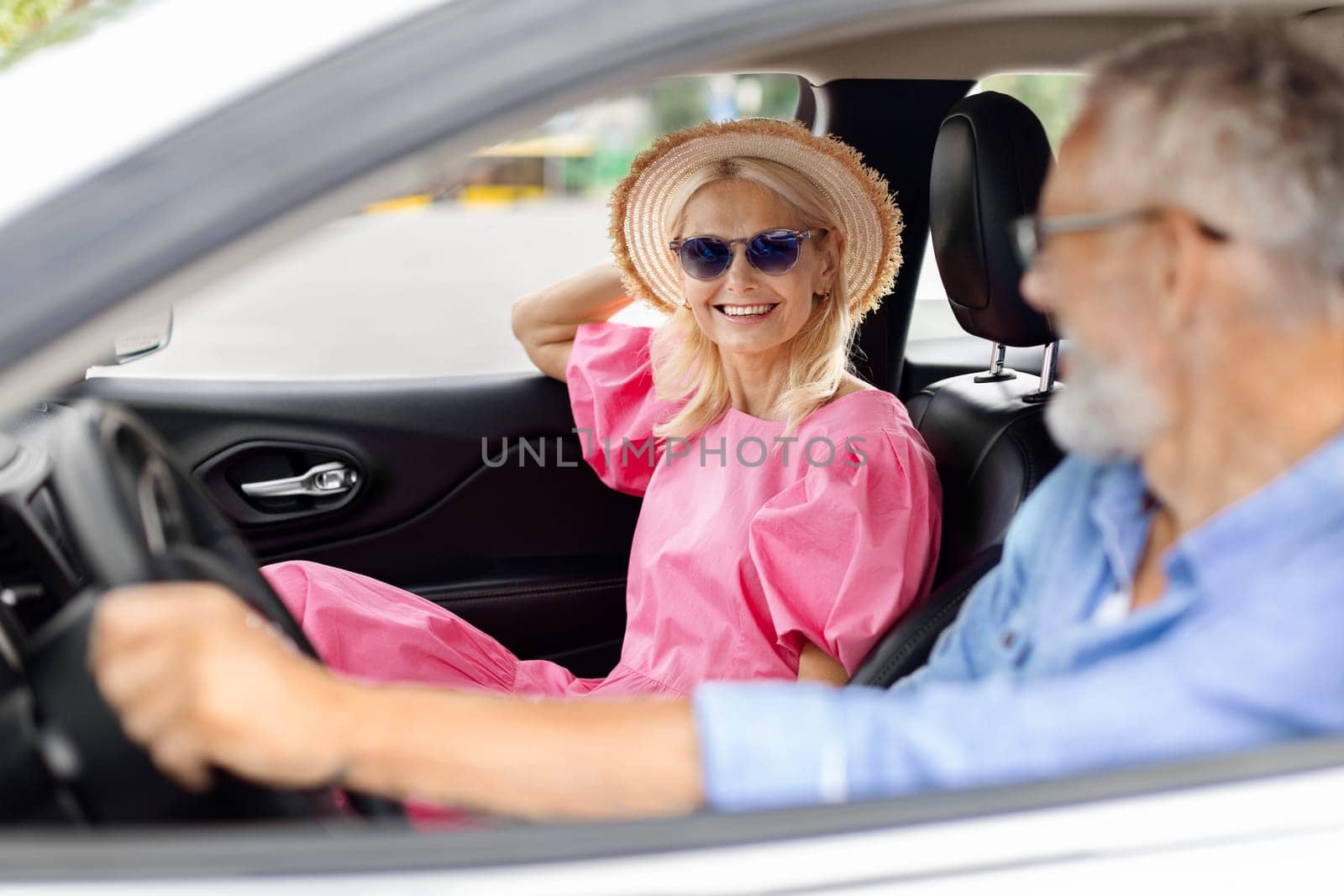 Smiling excited senior couple husband and wife going summer vacation by car together. Happy retired man and woman wearing wicker hat and sunglasses sitting in auto, enjoying road trip, chatting