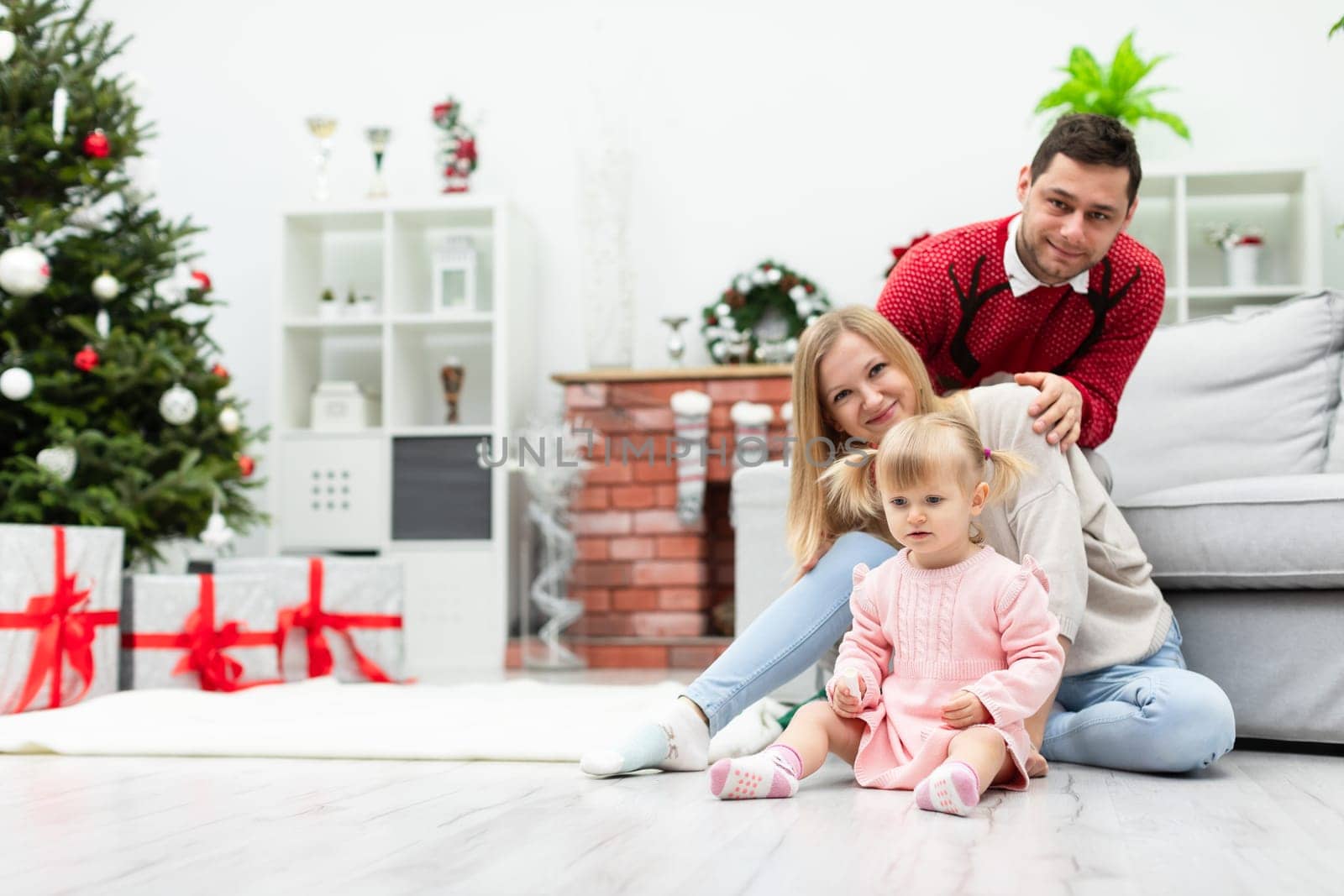 Parents with their little daughter are sitting on the floor in a bright room. In the background you can see a brick fireplace, lots of Christmas decorations and a Christmas tree. Under the Christmas tree stand large boxes of gifts.