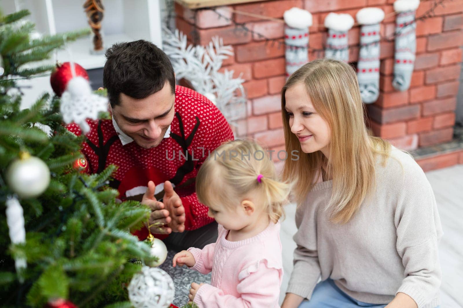 A woman, a man and a little girl are staying by the Christmas tree. The little girl is standing and her parents are crouching next to her. A man in a red sweater claps his hands. The family is smiling.
