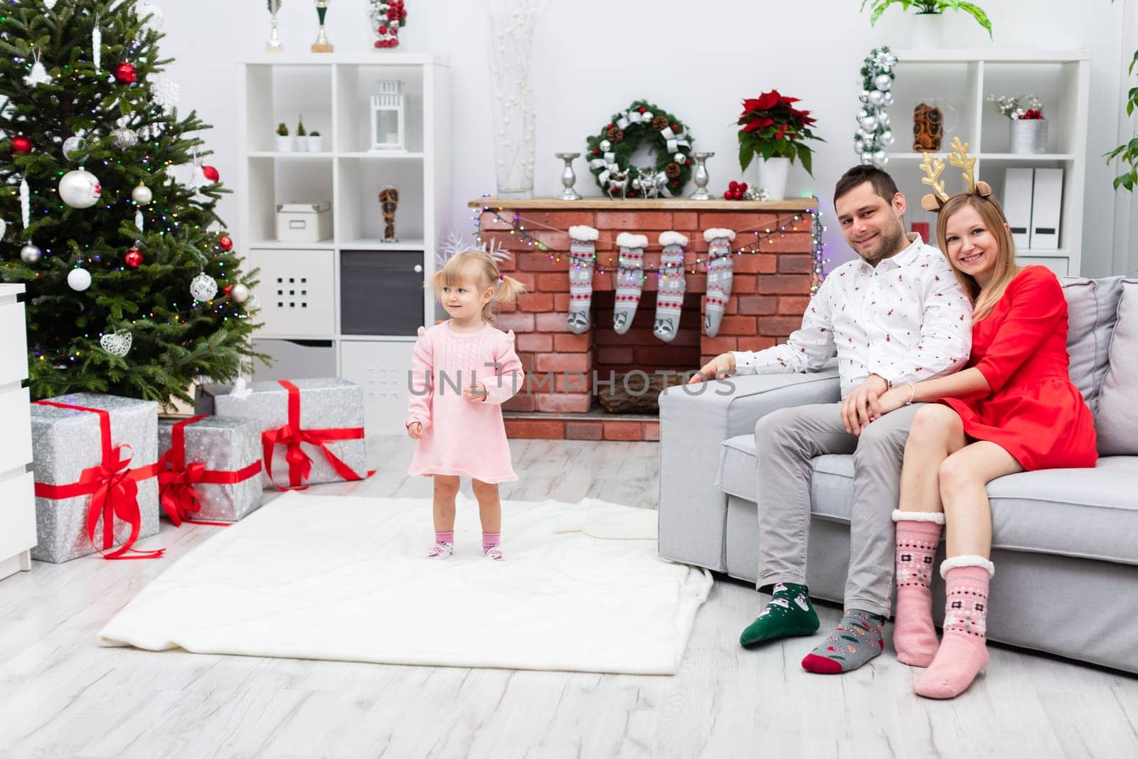 A hugging couple sits on a gray couch, while a little girl stands next to them. The girl stands on a white fluffy carpet. The family of three is staying in a room decorated festively.