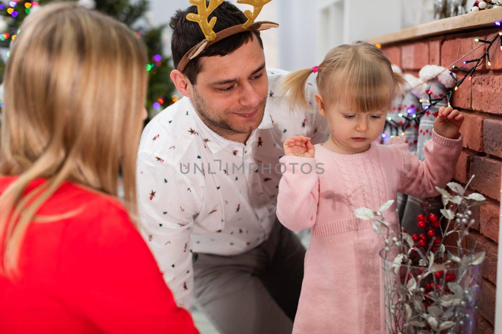 The man is wearing a headband with reindeer ears on his head. A woman, a man and a little girl are sitting together by the fireplace. The family is dressed in Christmas costumes. The little girl is watching the Christmas lights.