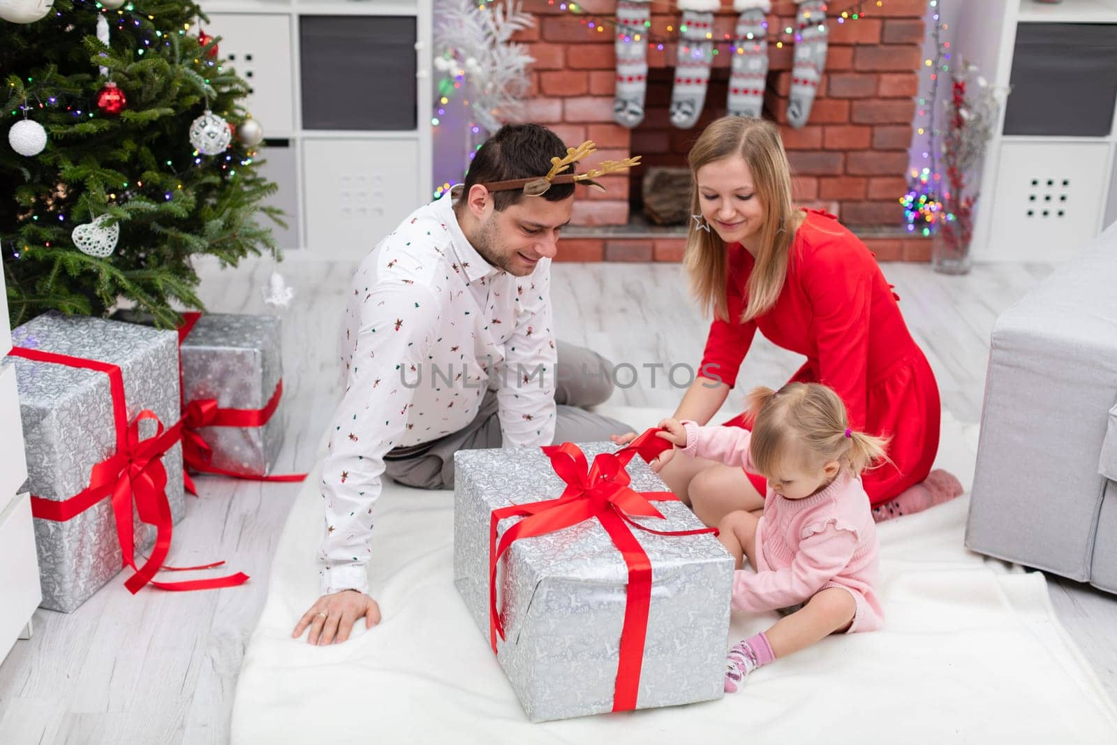 The woman is dressed in a red dress. A woman, a man and a little girl are sitting on the floor. The family is sitting around a large Christmas present. A section of a Christmas tree can be seen next to the family.