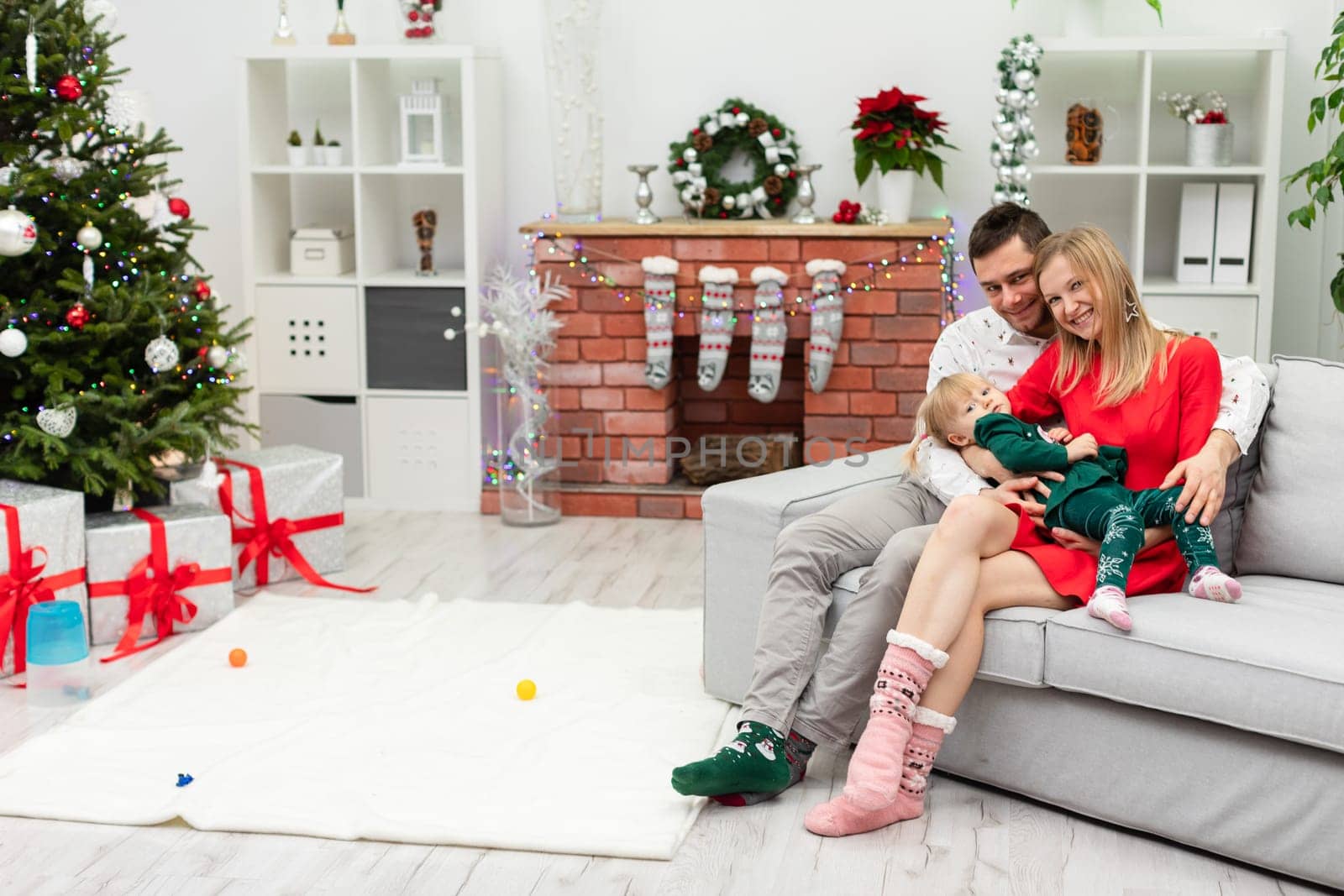 A woman wearing a red dress holds a little girl on her lap. The child is wearing a dark green tracksuit. A man sits next to the woman and the little girl and embraces his wife and daughter.