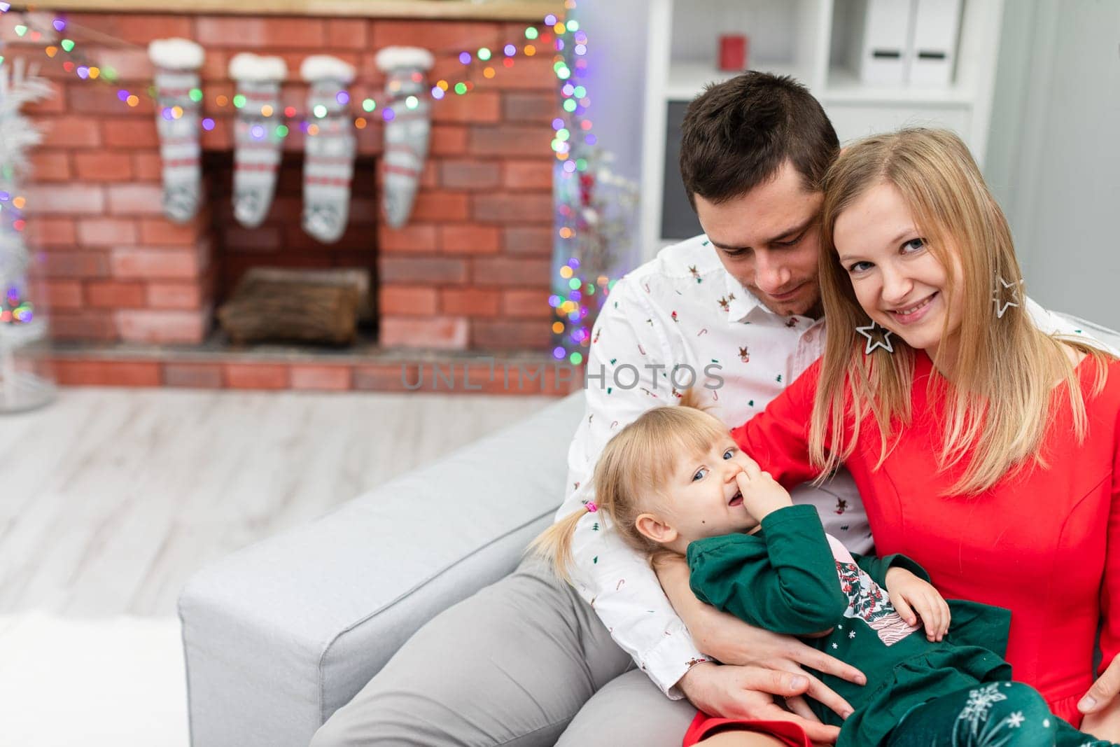 A man, a woman and a little girl sit together on a gray couch. The woman holds her lying daughter in her lap. The blurry background shows a brick fireplace decorated with Christmas lights.