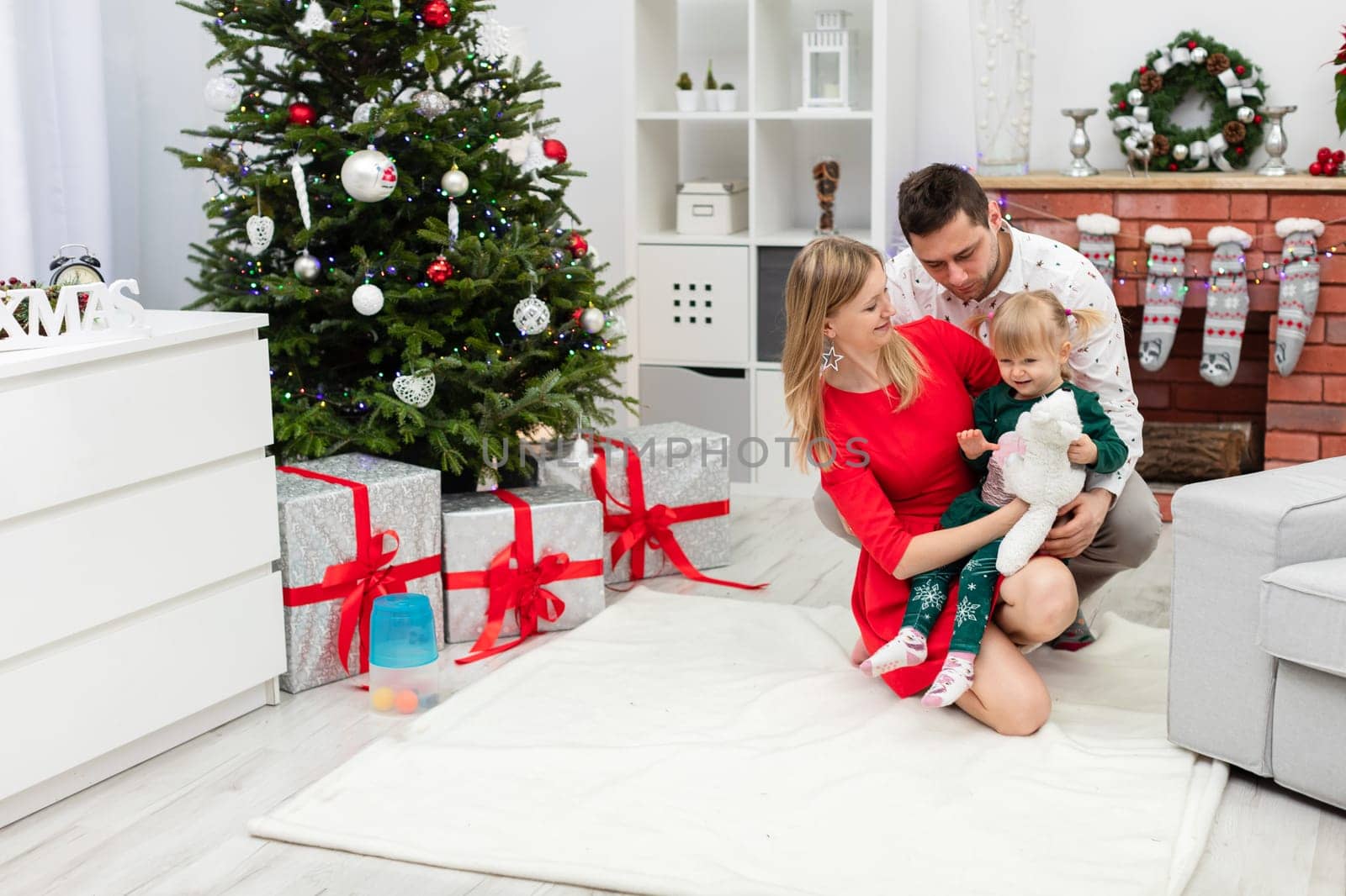 Under the decorated Christmas tree lie large gifts tied with red ribbons. Next to the Christmas tree is a man, a woman and a little girl. The mother is hugging her little daughter. The family members smile at each other.