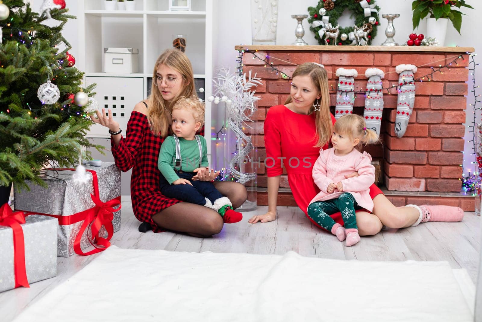 Two women are sitting by the fireplace. Each of them is holding a small child on her lap. One of the women turns her back and looks at the baubles hanging on the Christmas tree. The room is decorated with Christmas decorations.