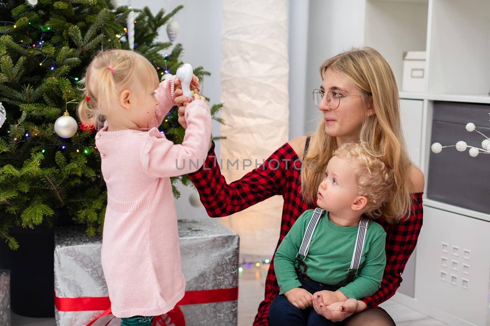 In a room decorated with Christmas ornaments there is a woman and two young children. The woman is playing with a boy and a girl. The woman has blonde hair and glasses. They are all sitting by the Christmas tree.