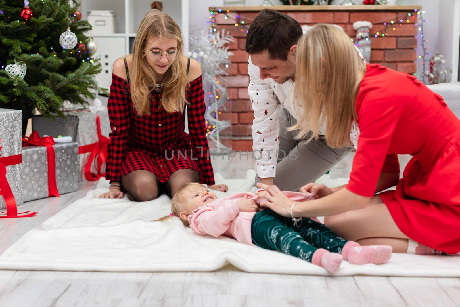 A woman in a red dress, a man in a white shirt and a woman in a plaid dress are sitting on a white carpet. Next to them lies a little girl and laughs while playing.