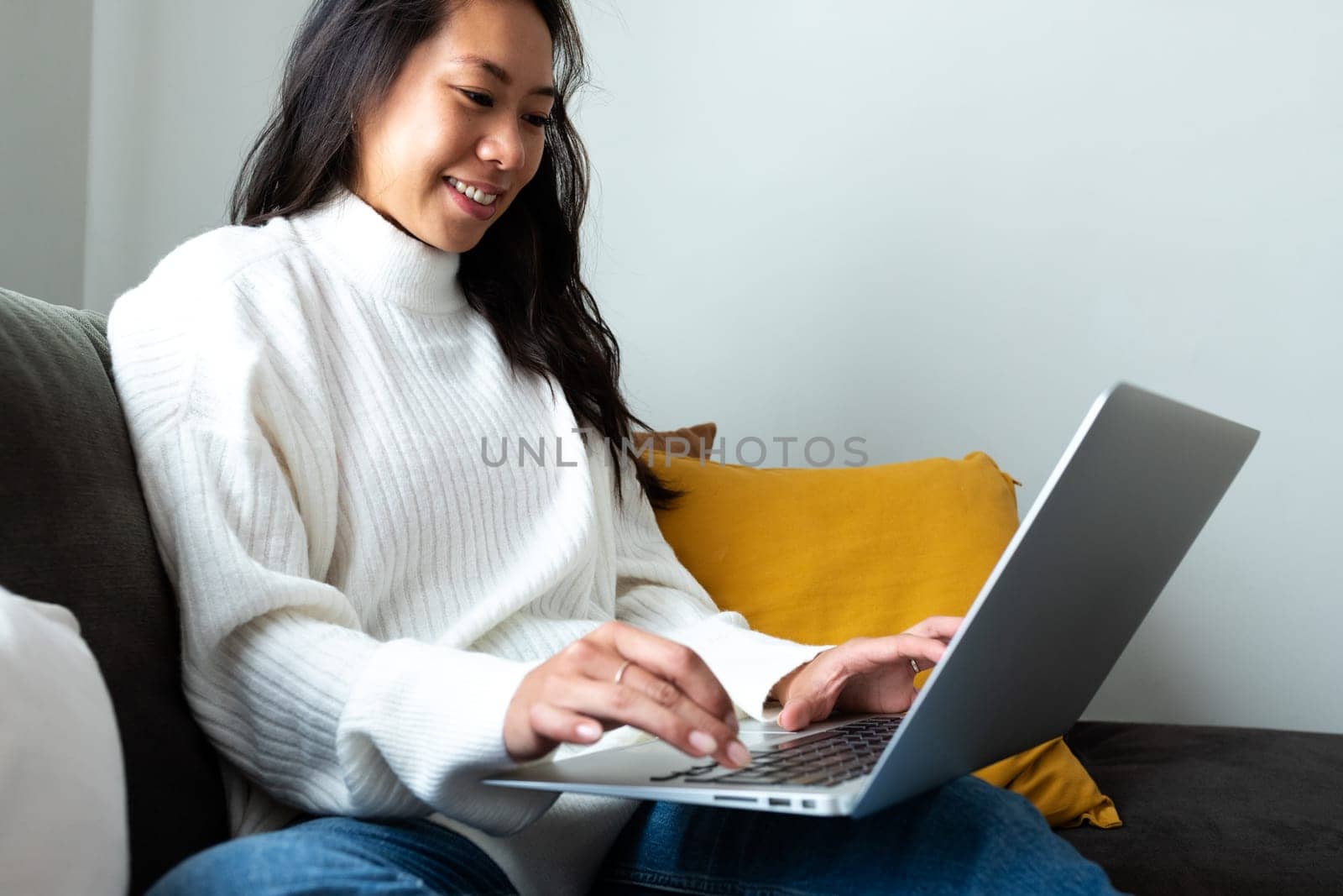 Young Asian woman sitting on the sofa using laptop working at home living room. by Hoverstock