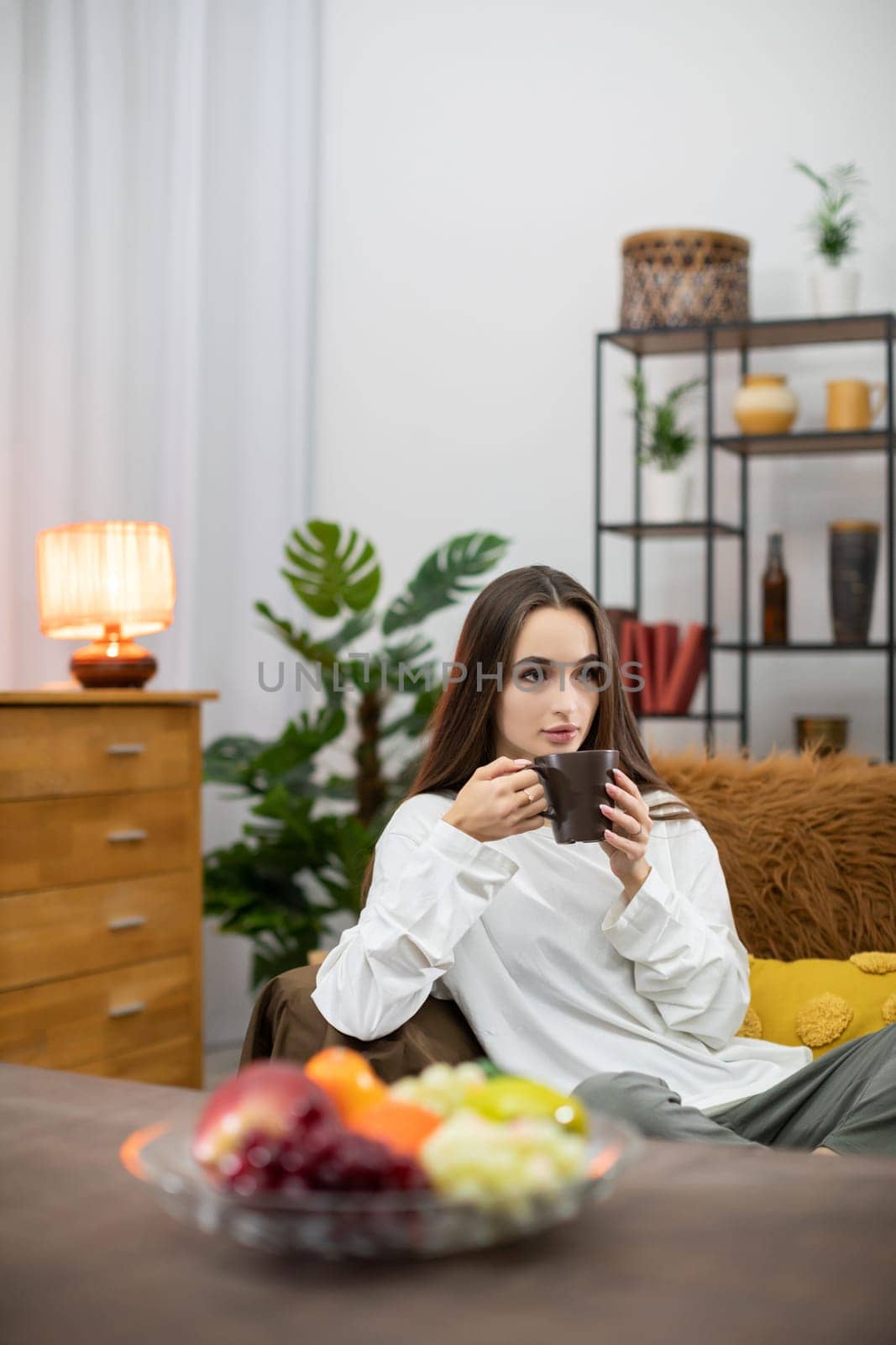 A young woman relaxes with tea in a cozy living room filled with plants and fresh fruits around her