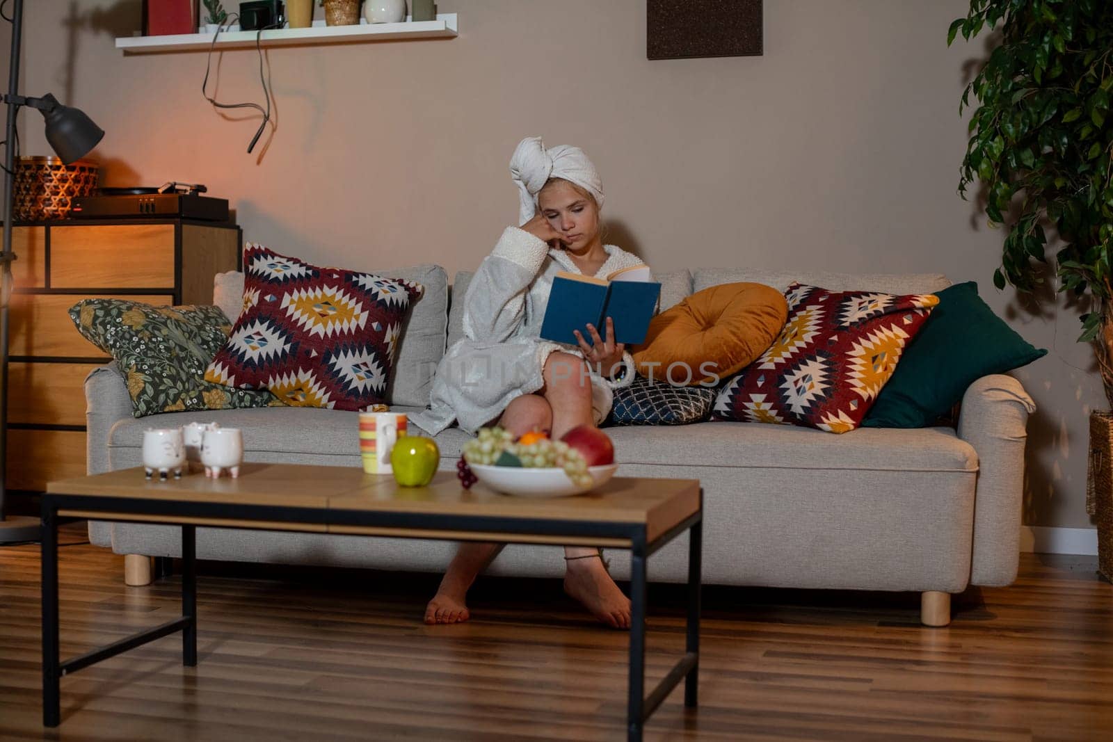 A woman enjoys a cozy moment reading her favorite book on the sofa, with snacks and a refreshing drink
