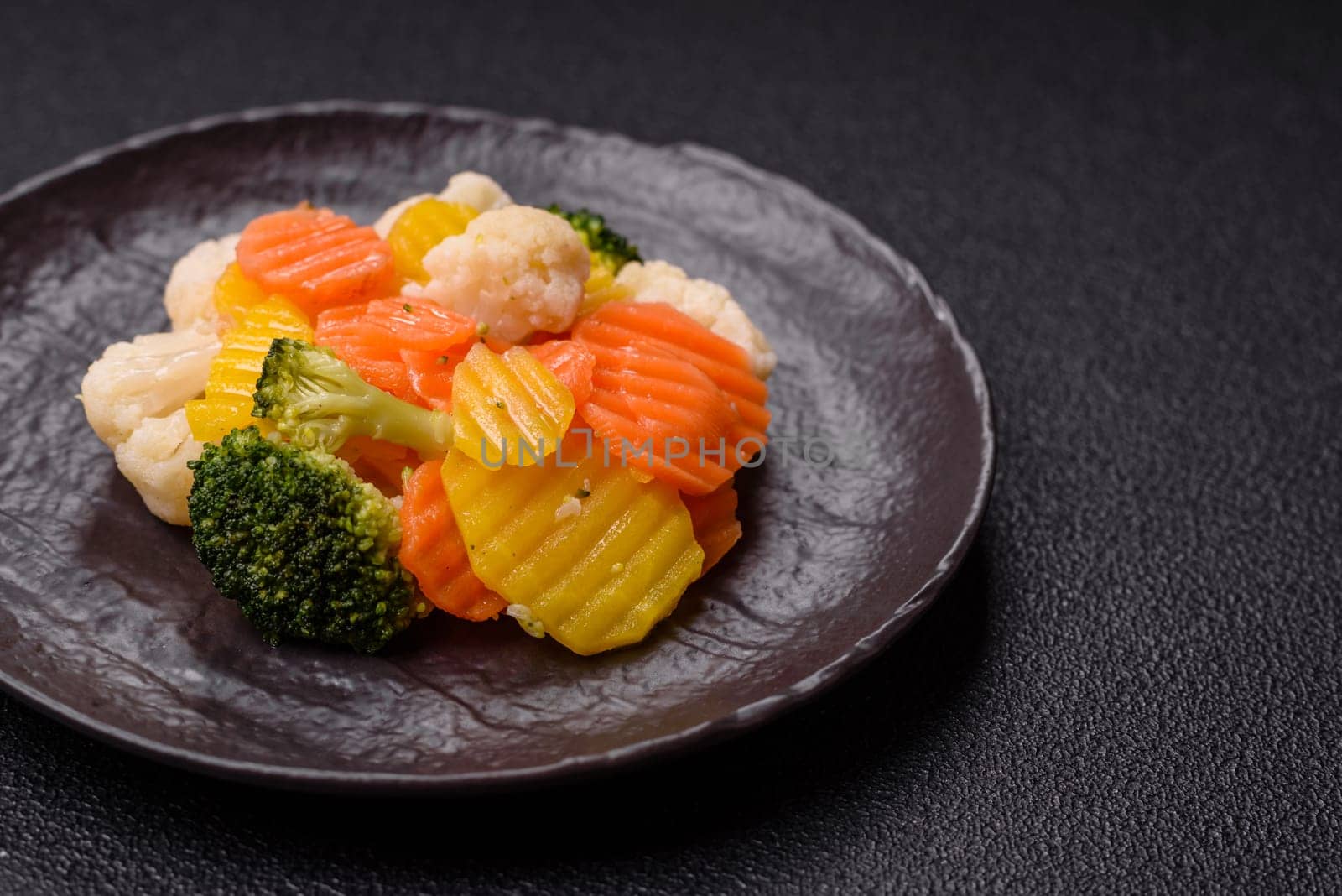 Delicious fresh vegetables steamed carrots, broccoli, cauliflower on a black plate on a dark concrete background
