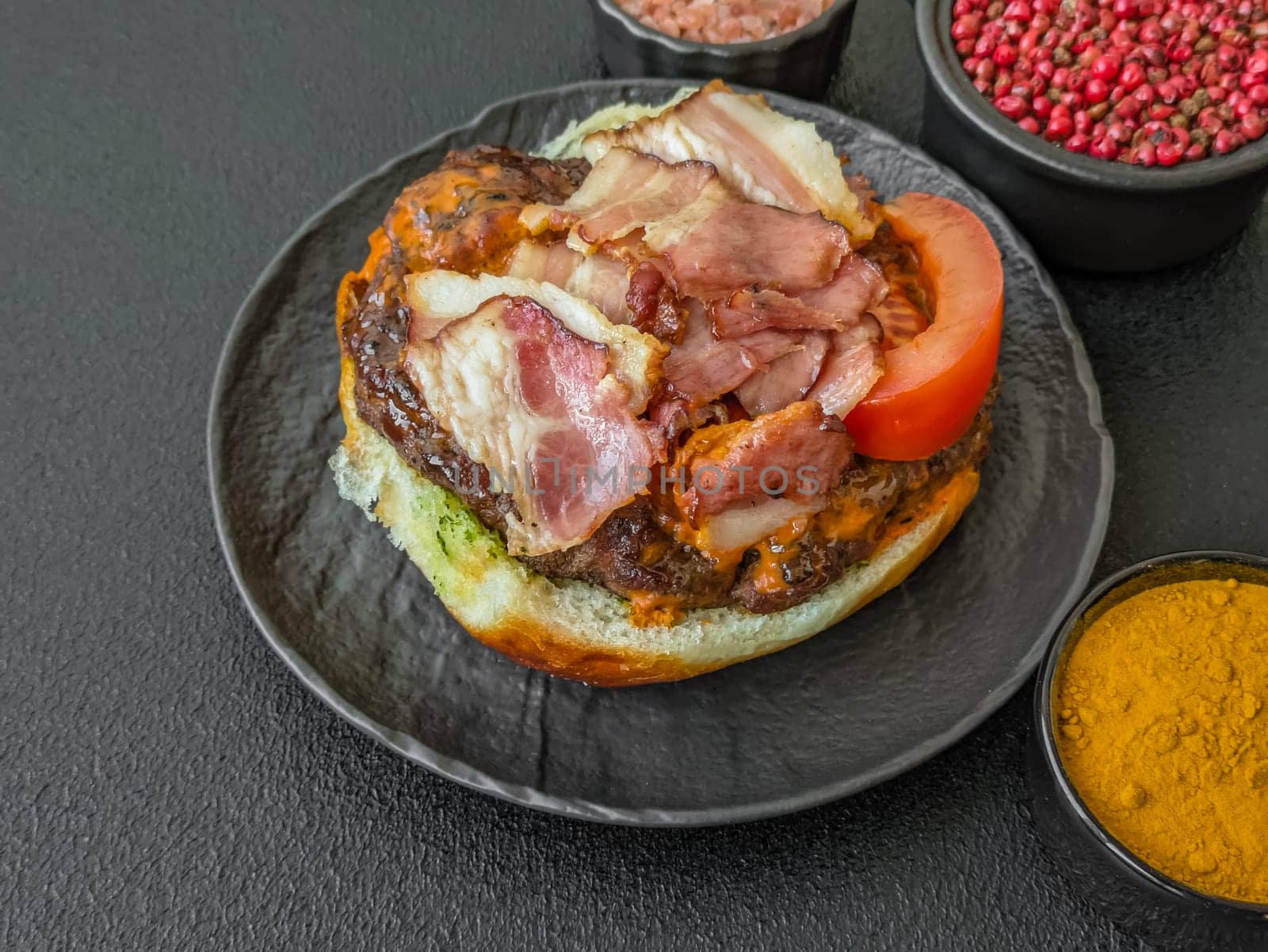 Juicy burger with beef patty, tomatoes, cheese, bacon, salt and spices on dark concrete background