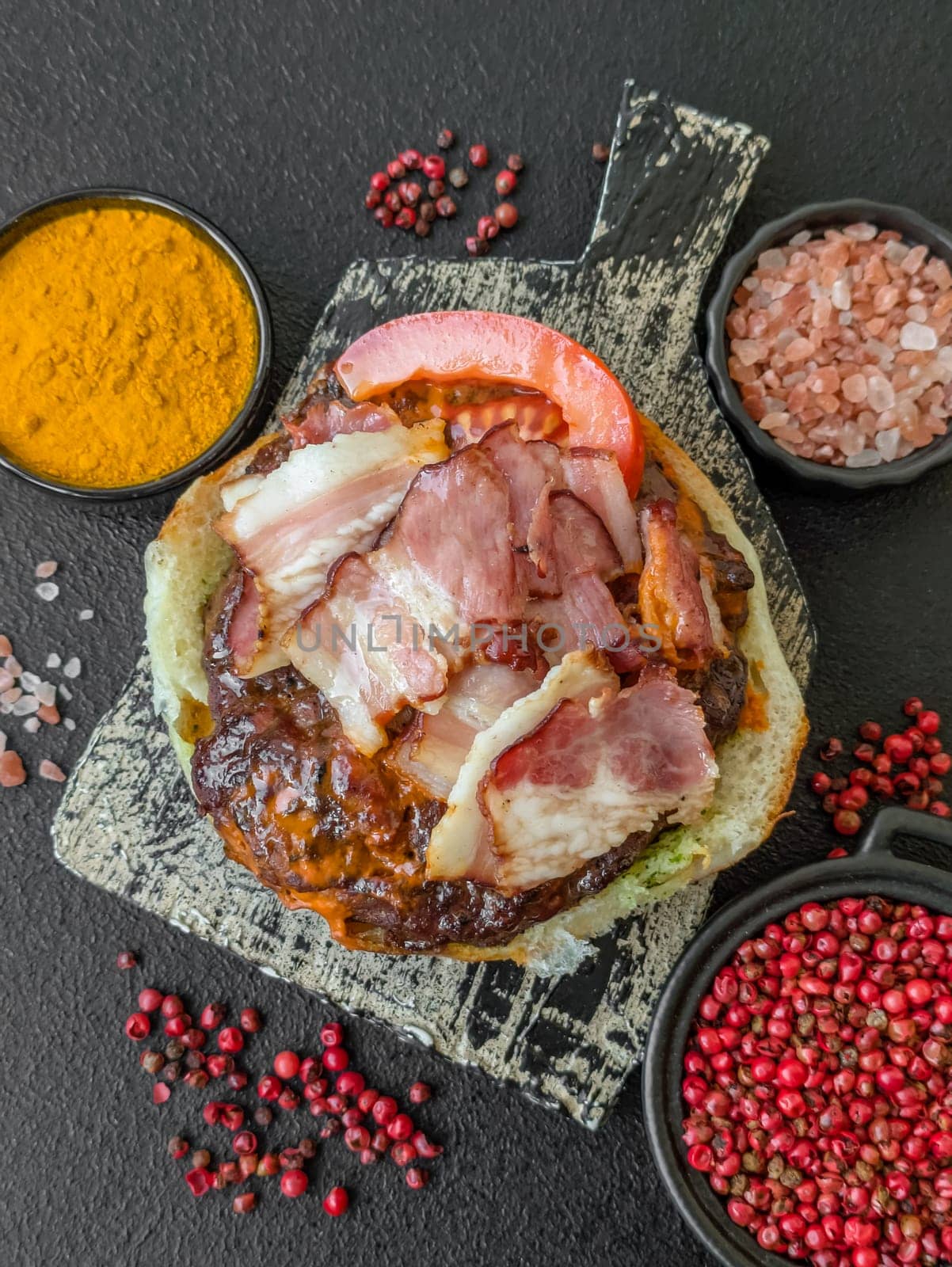 Juicy burger with beef patty, tomatoes, cheese, bacon, salt and spices on dark concrete background