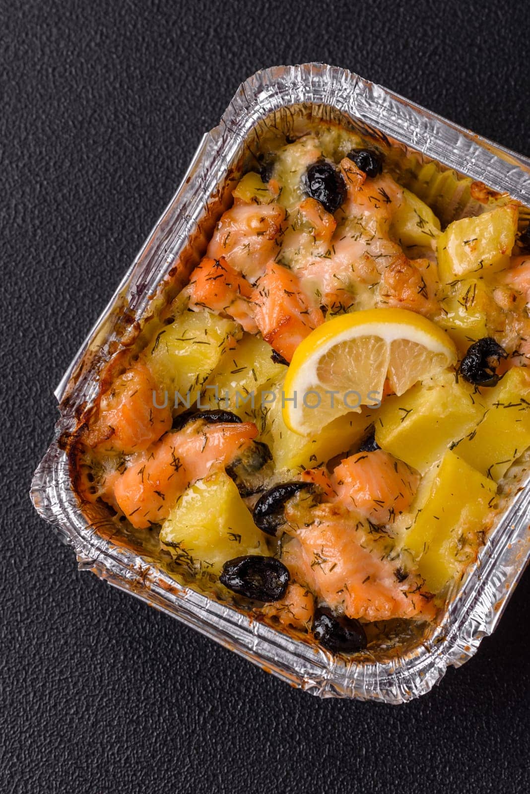 Baked salmon pieces with potatoes, cheese and olives with salt, spices and herbs on a dark concrete background