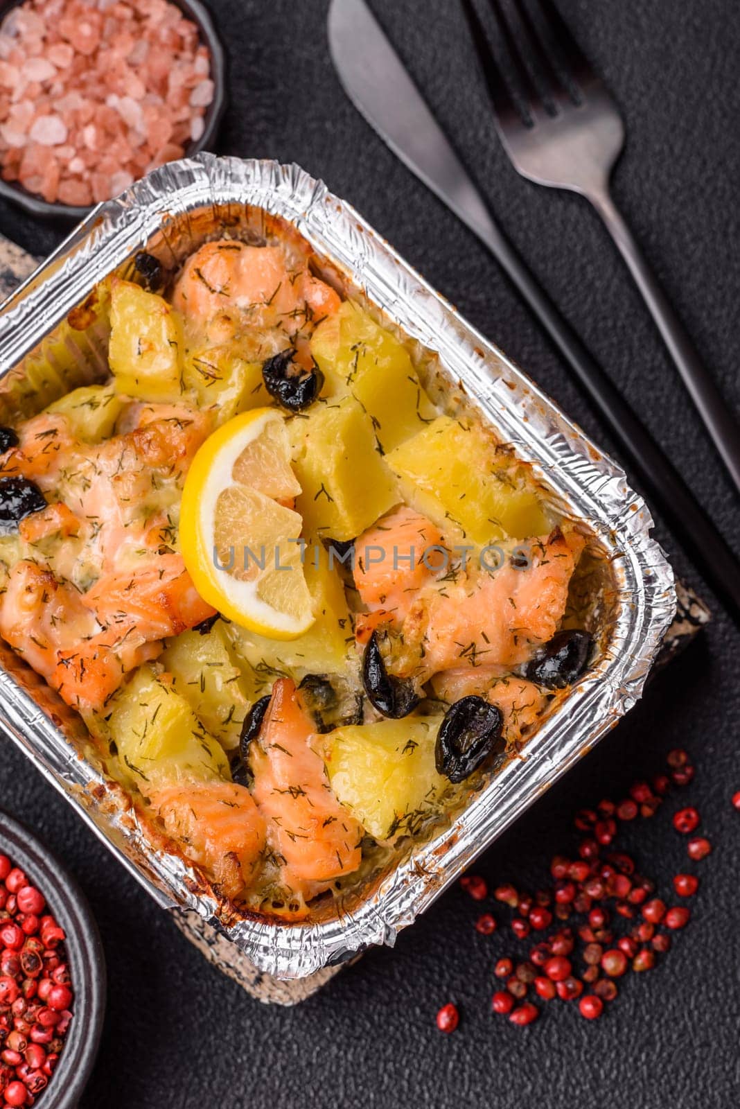 Baked salmon pieces with potatoes, cheese and olives with salt, spices and herbs on a dark concrete background