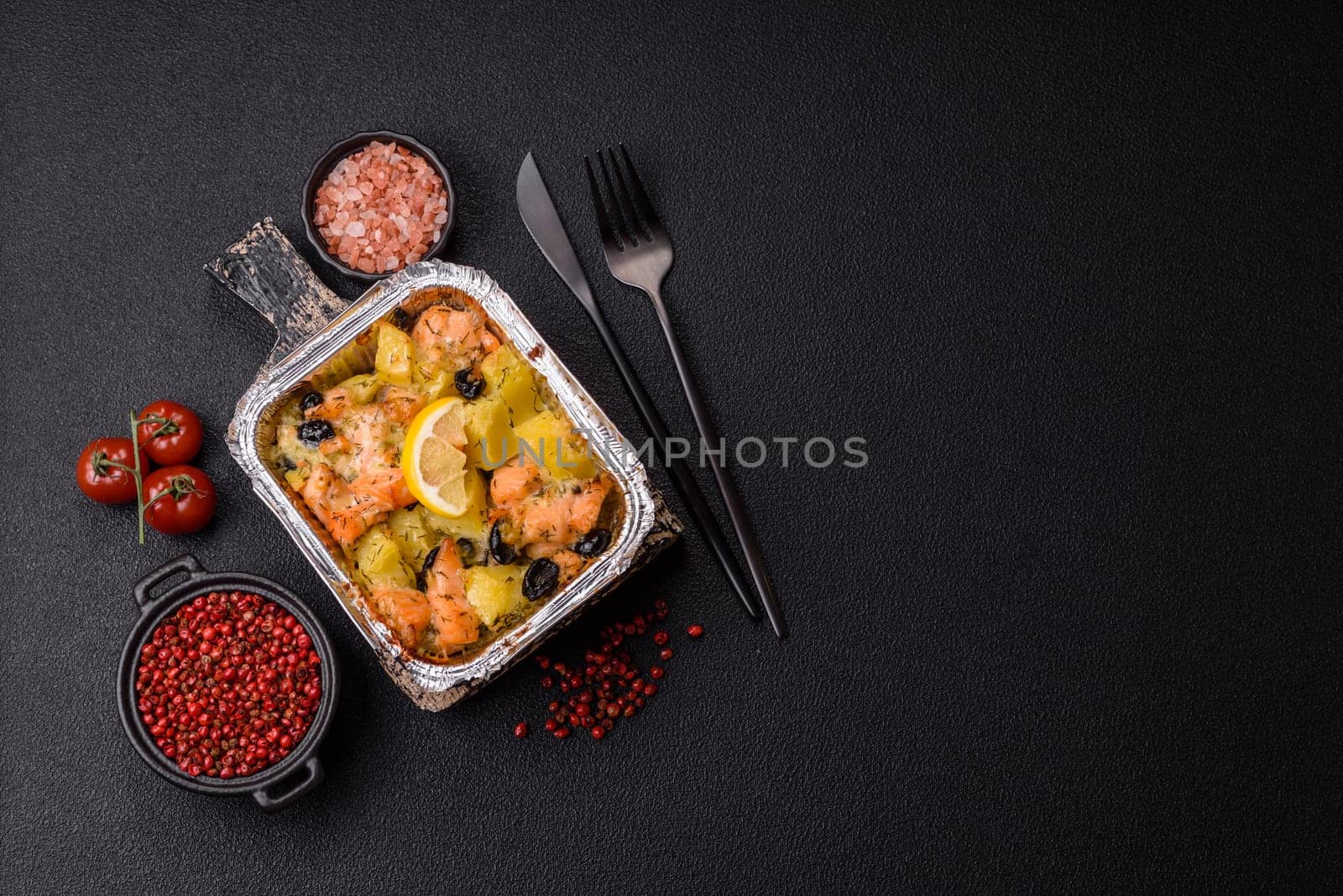 Baked salmon pieces with potatoes, cheese and olives with salt, spices and herbs on a dark concrete background