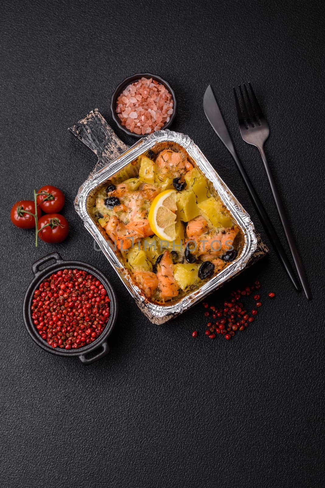 Baked salmon pieces with potatoes, cheese and olives with salt, spices and herbs on a dark concrete background