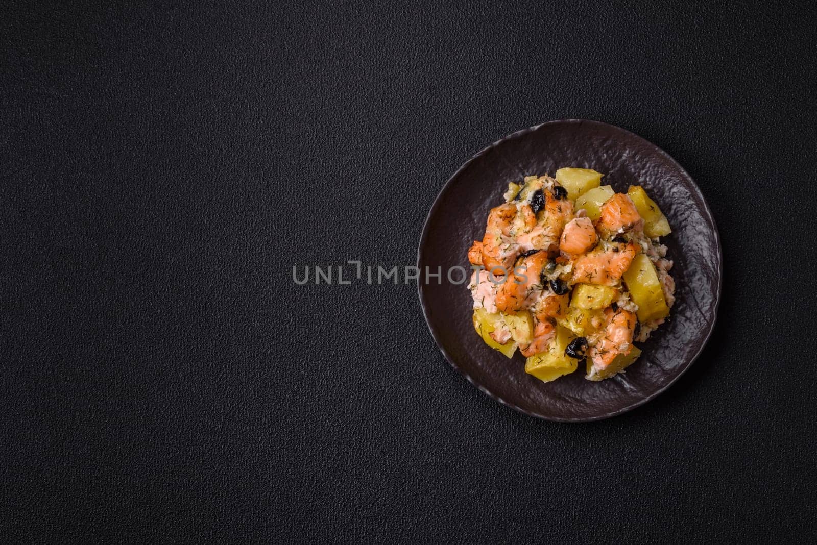 Baked salmon pieces with potatoes, cheese and olives with salt, spices and herbs on a dark concrete background