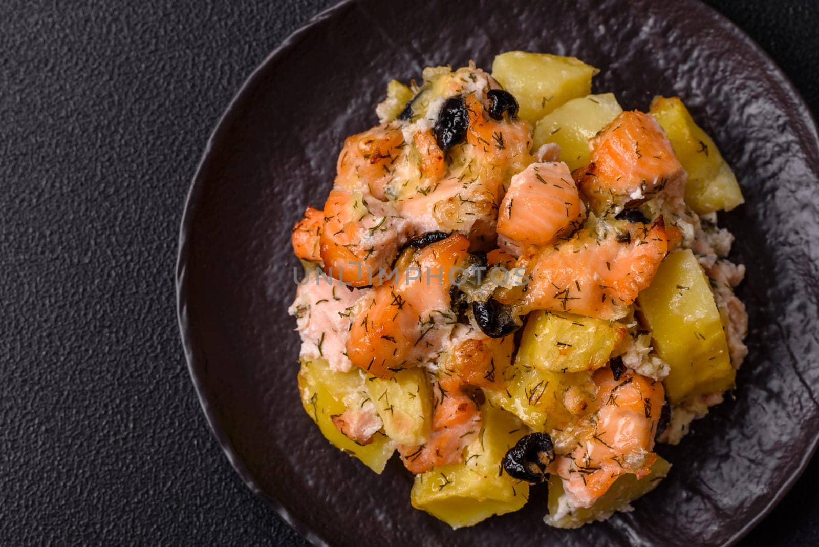 Baked salmon pieces with potatoes, cheese and olives with salt, spices and herbs on a dark concrete background