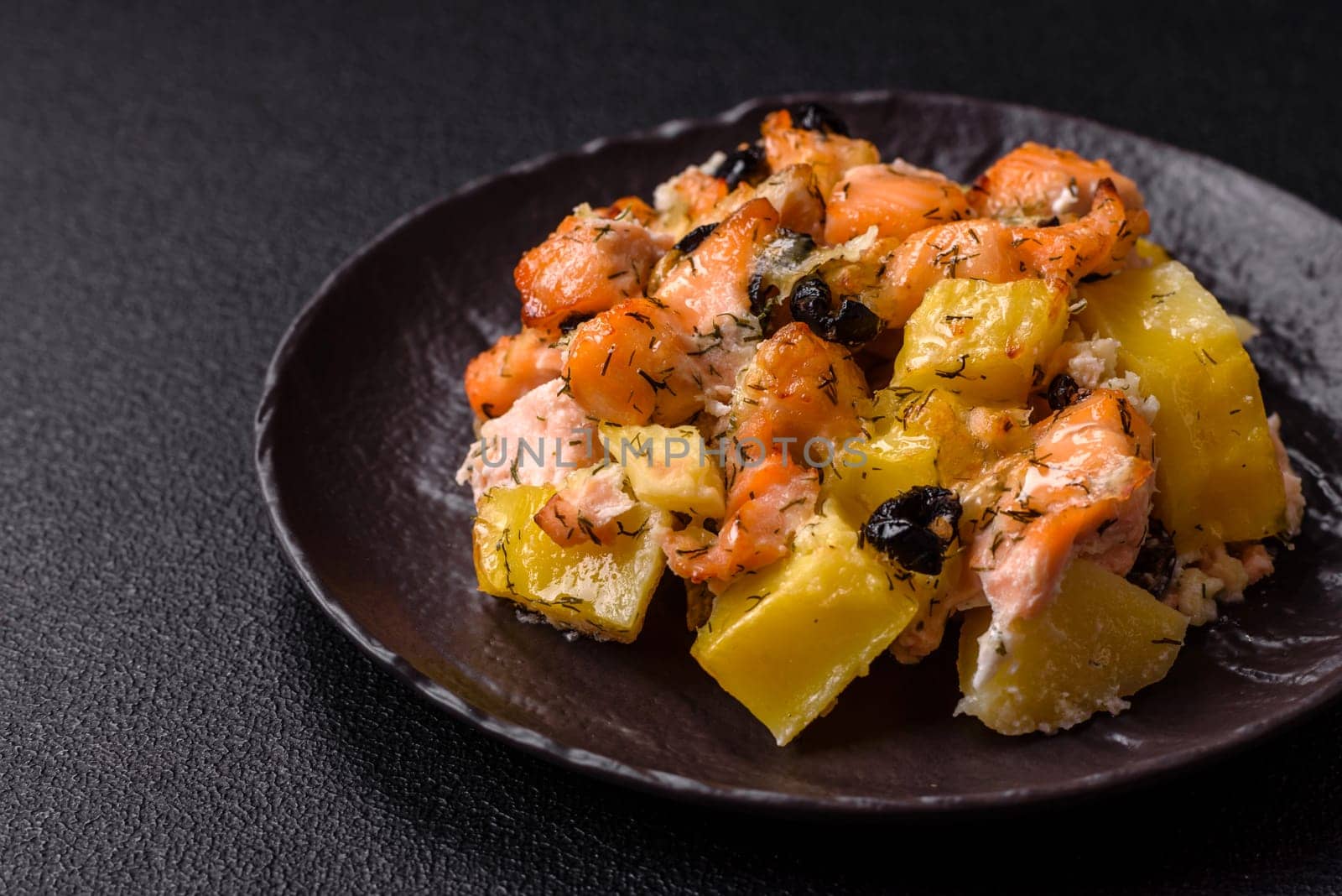 Baked salmon pieces with potatoes, cheese and olives with salt, spices and herbs on a dark concrete background