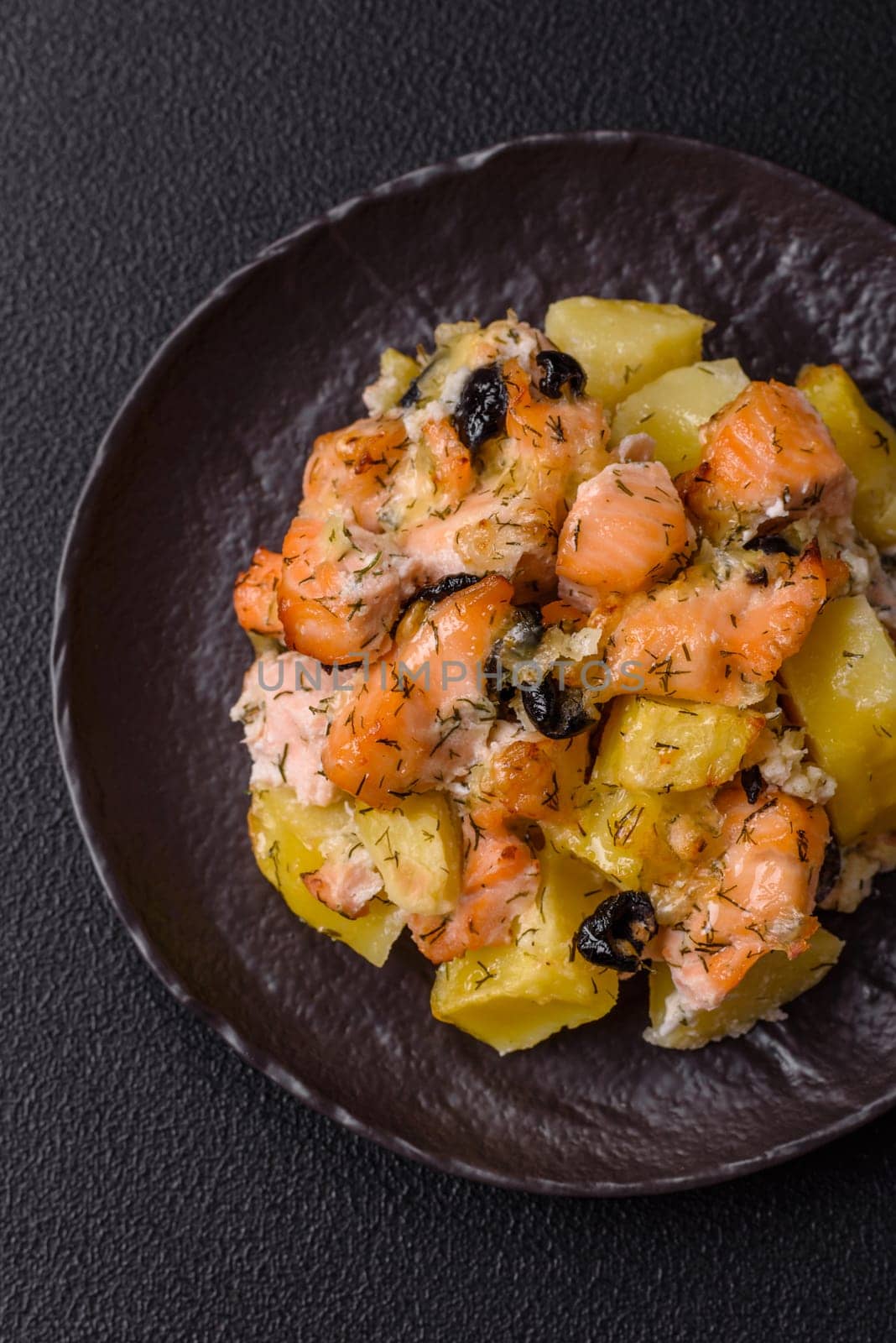 Baked salmon pieces with potatoes, cheese and olives with salt, spices and herbs on a dark concrete background