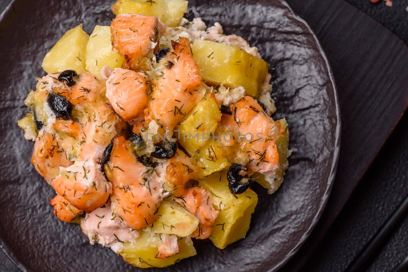 Baked salmon pieces with potatoes, cheese and olives with salt, spices and herbs on a dark concrete background