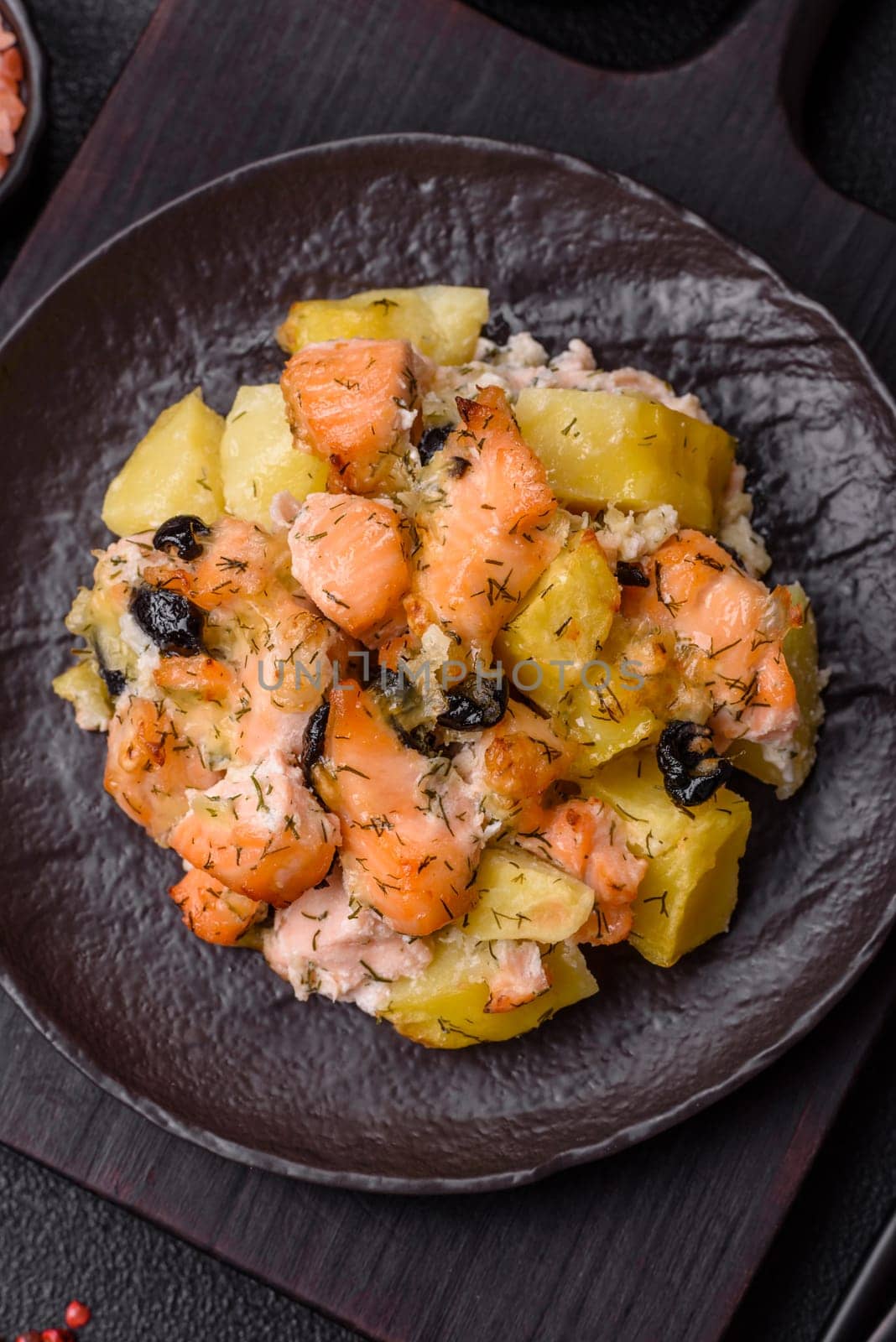 Baked salmon pieces with potatoes, cheese and olives with salt, spices and herbs on a dark concrete background