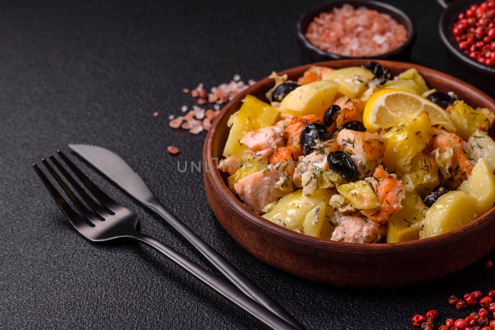 Baked salmon pieces with potatoes, cheese and olives with salt, spices and herbs on a dark concrete background