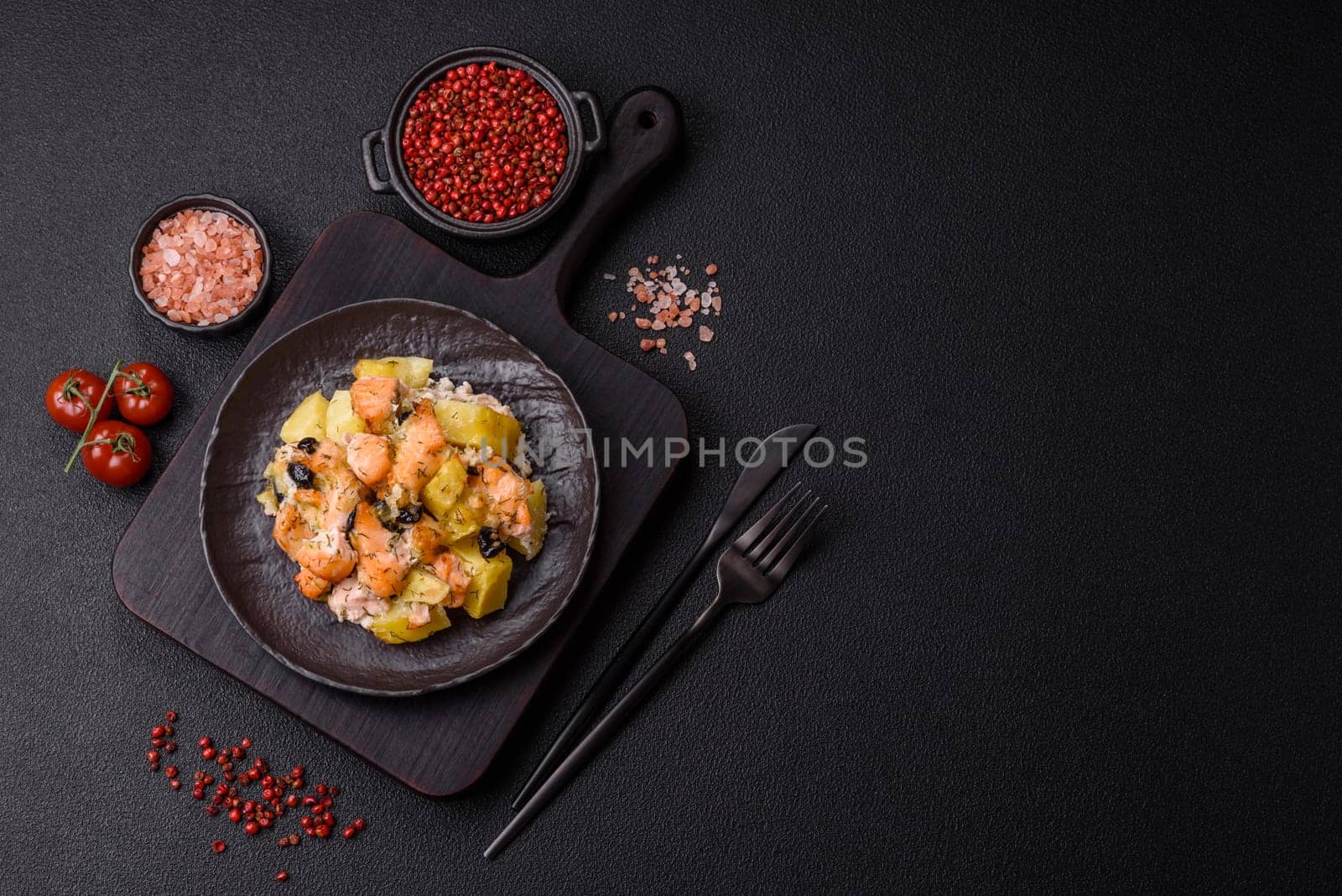 Baked salmon pieces with potatoes, cheese and olives with salt, spices and herbs on a dark concrete background