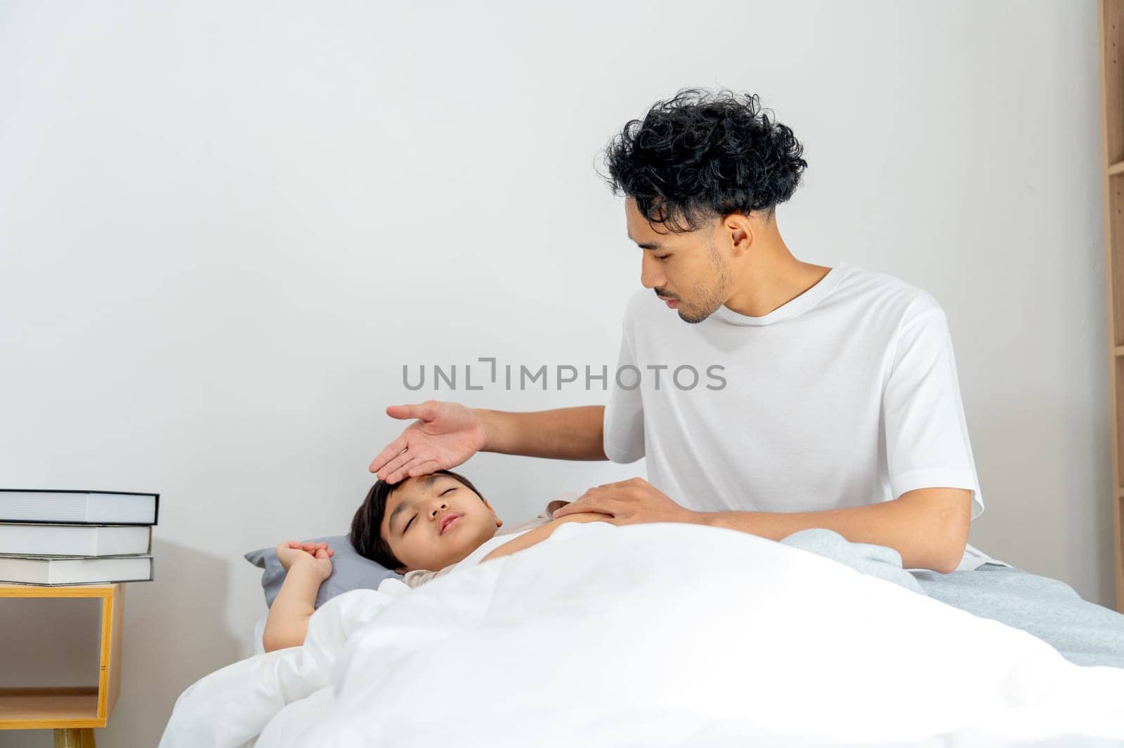 Front view Asian young father sit near his son lie on bed with sick or unwell and father take care with touch son forehead to check temperature.