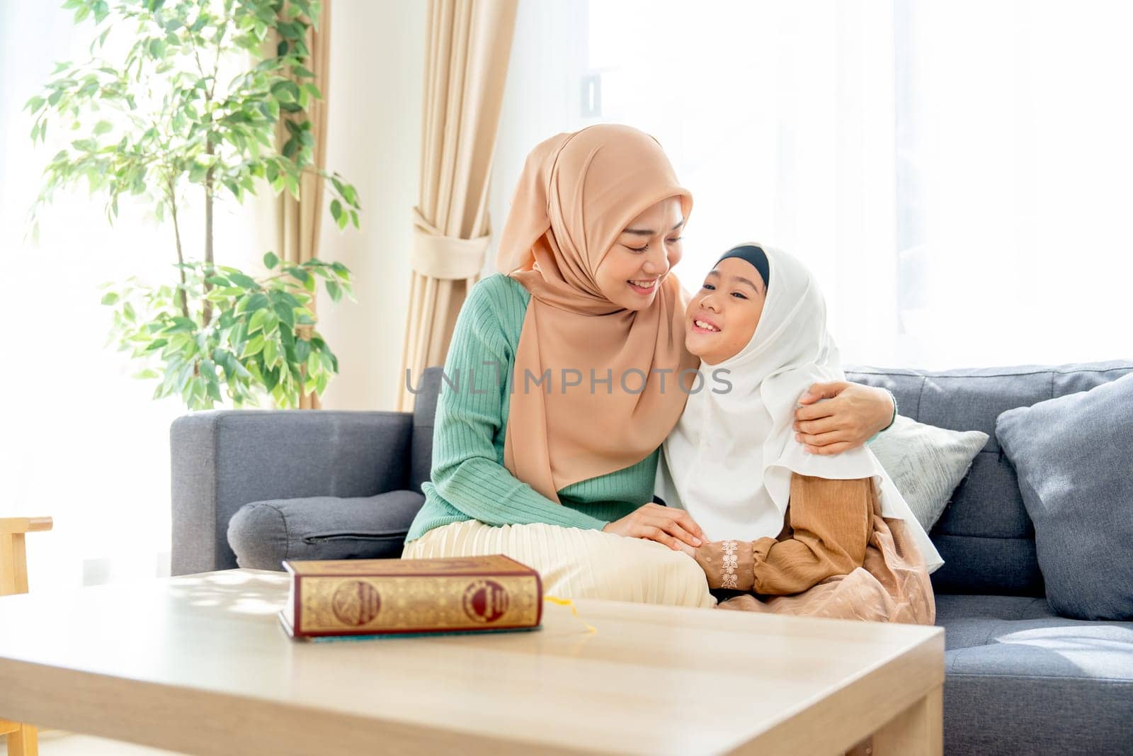 Wide shot of Asian muslim mother sit on sofa beside of her daughter and hug her child they also look face each other with smiling in concept of happiness muslim family at home.