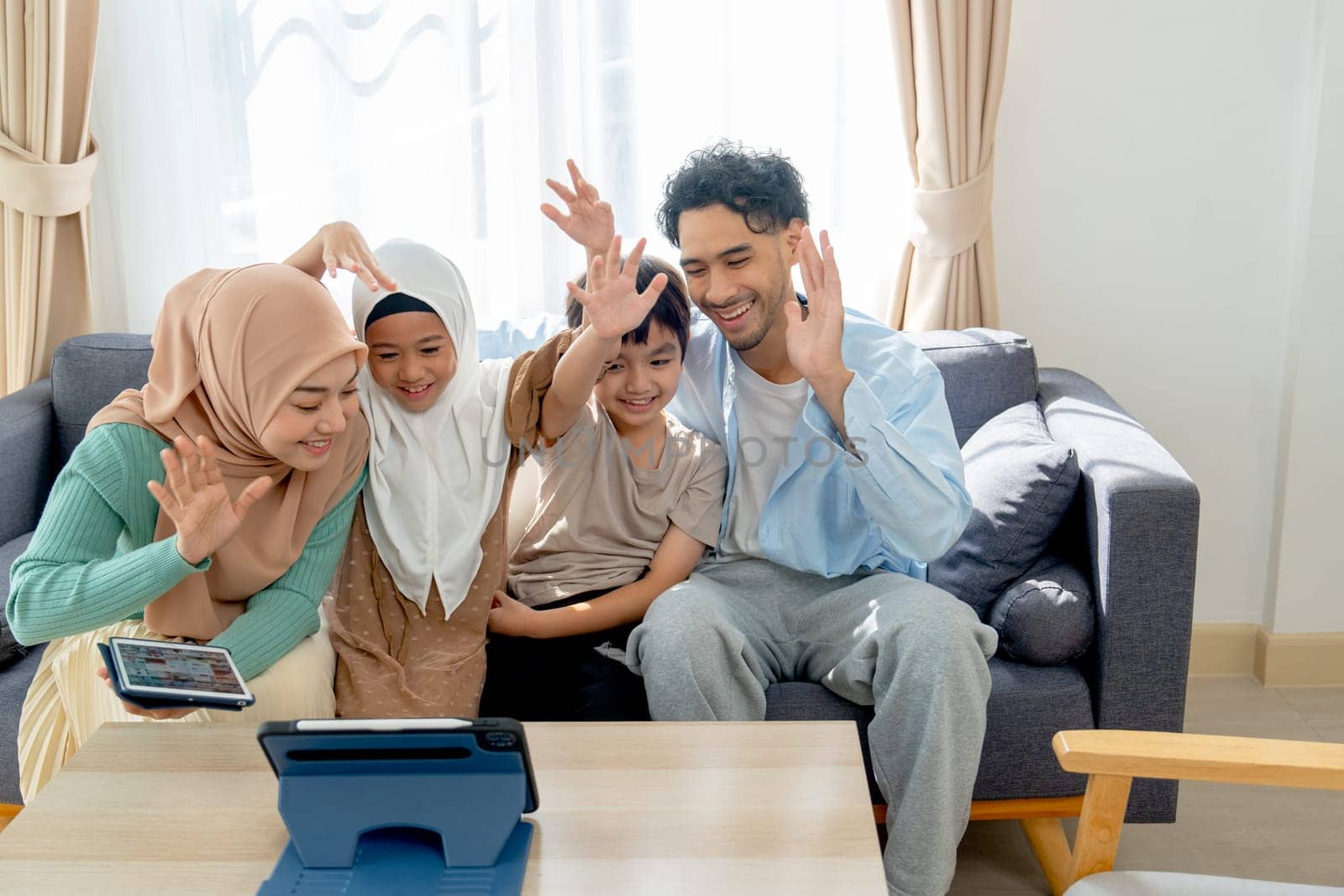 Front view of Asian muslim family compose of young mother, father, daughter and son hug and stay together on sofa in living room of their house and show greeting or say hi to tablet for online meeting