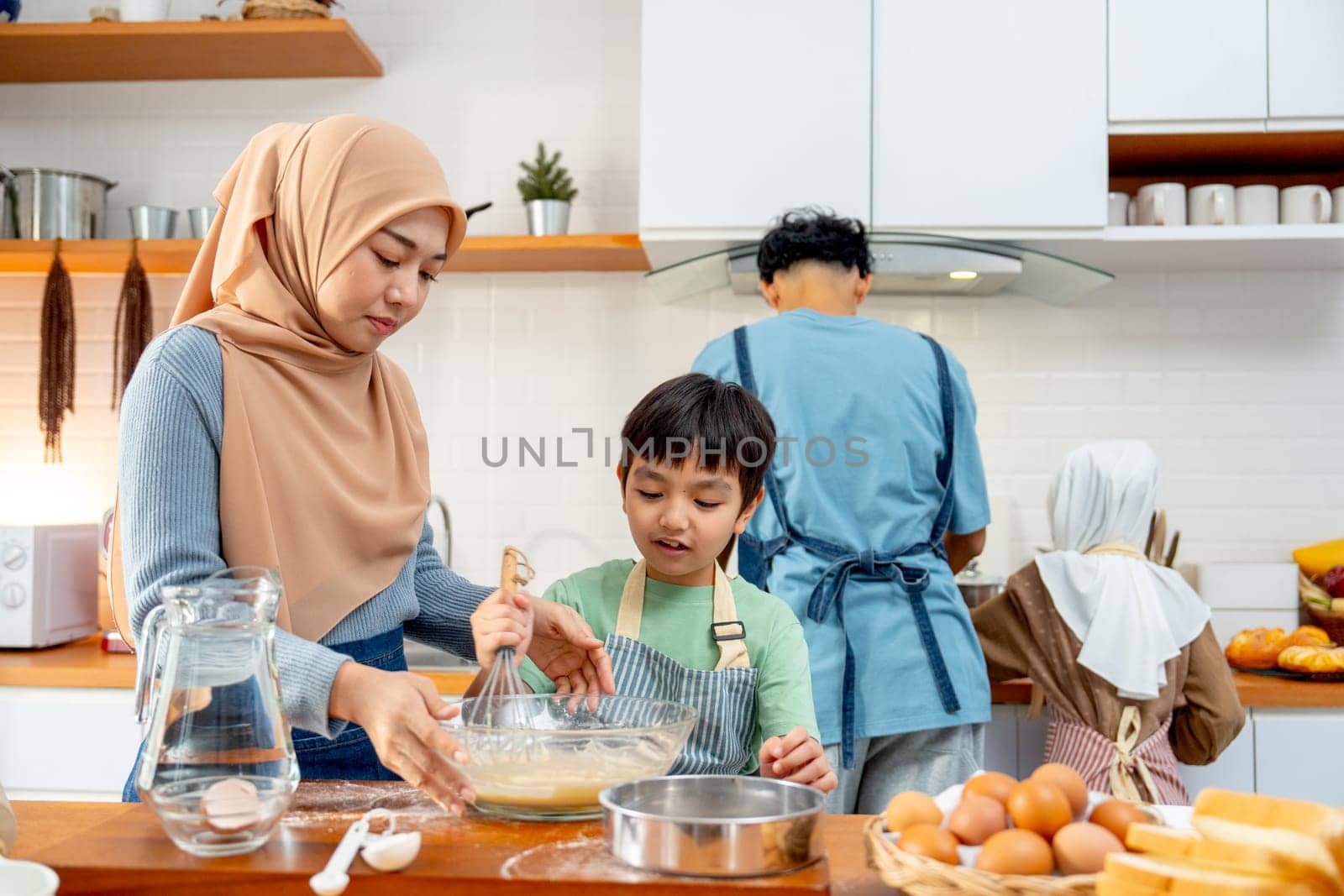 Muslim mother stand beside of her son to mix flower in glass bowl and the other also cook in the back of kitchen in their house.