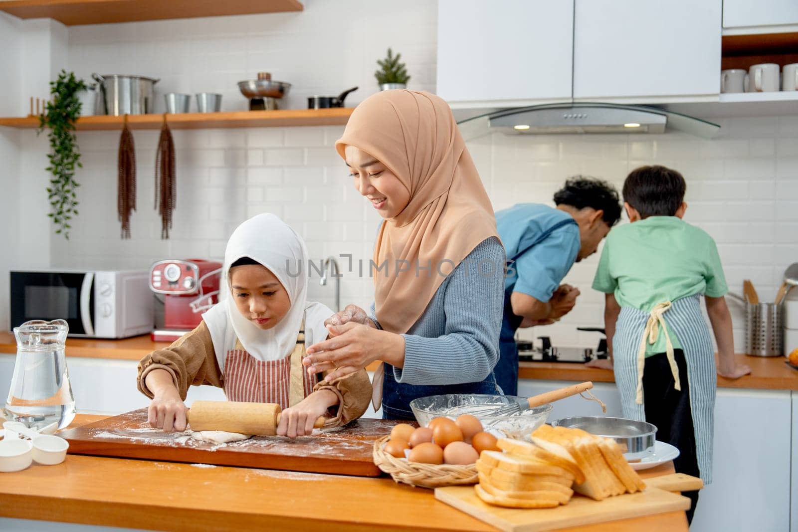 Wide shot of little Asian muslim girl enjoy to use rolling pin to make some bakery and support beside by her young mother in the kitchen with father and brother also cooking in the back.