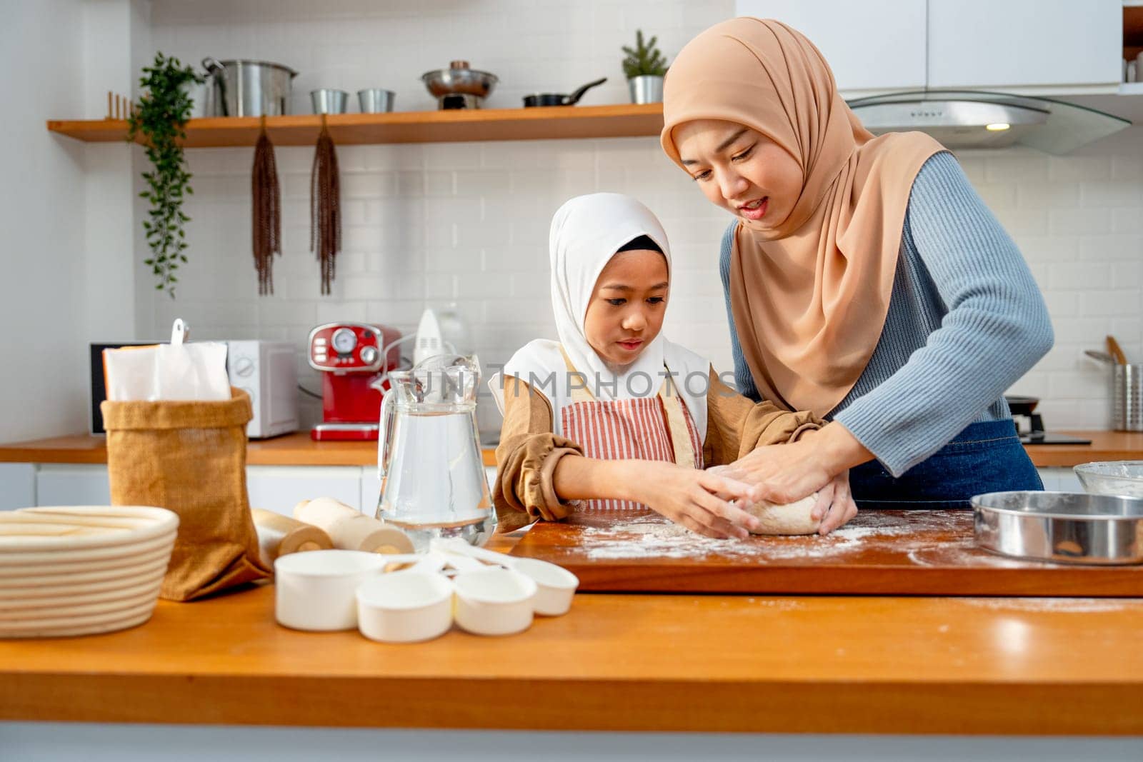 Young Asian muslim mother help and teach her daughter to threshing flour by hands on wooden tray for making the bakery in the kitchen of their house.