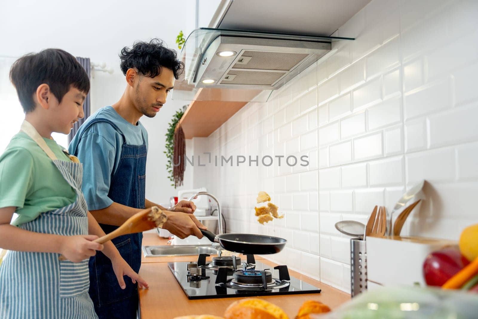 Young Asian father show skill of cooking by throwing pan cake up and his son cheer up beside in the kitchen of their house.